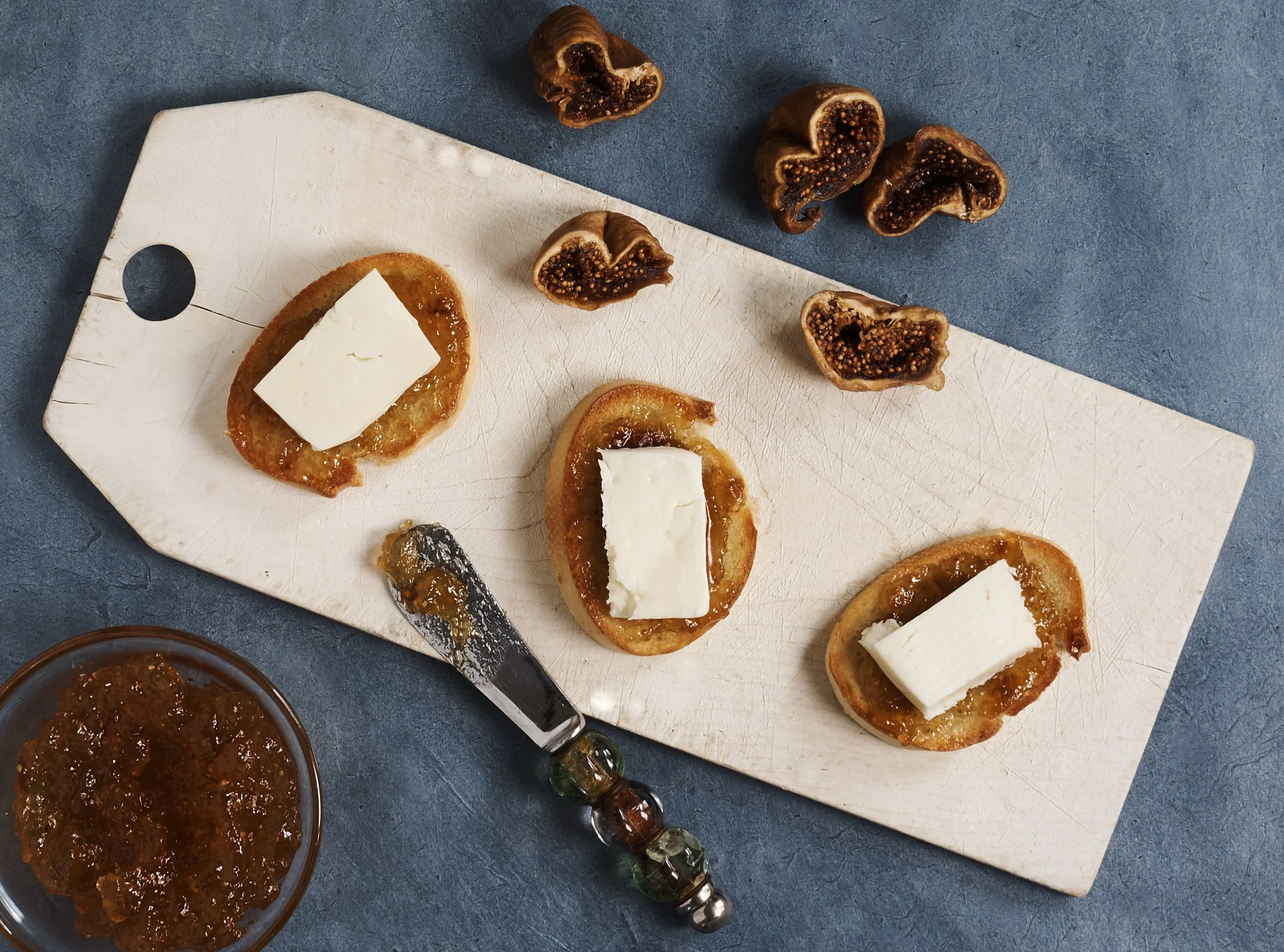 Several slices of toasted baguette topped with apricot jam and Kasbo's jibenh, syrian cheese, placed on a wooden cutting board. There are also halved figs with dark interiors, a knife with jam, and a small bowl of jam on a blue surface.