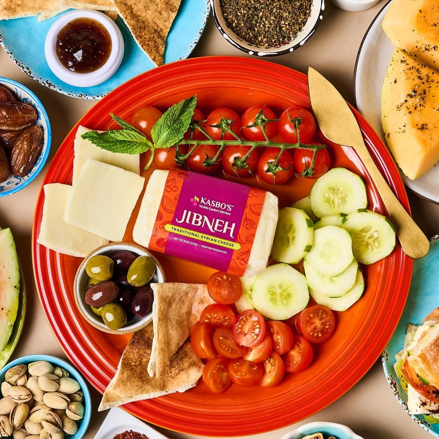 A colorful table setting with a large orange plate containing cherry tomatoes, sliced cucumbers, sliced tomatoes, and a block of Syrian cheese labeled Jibneh. Surrounding bowls contain olives, chopped pistachios, and various dips and herbs.