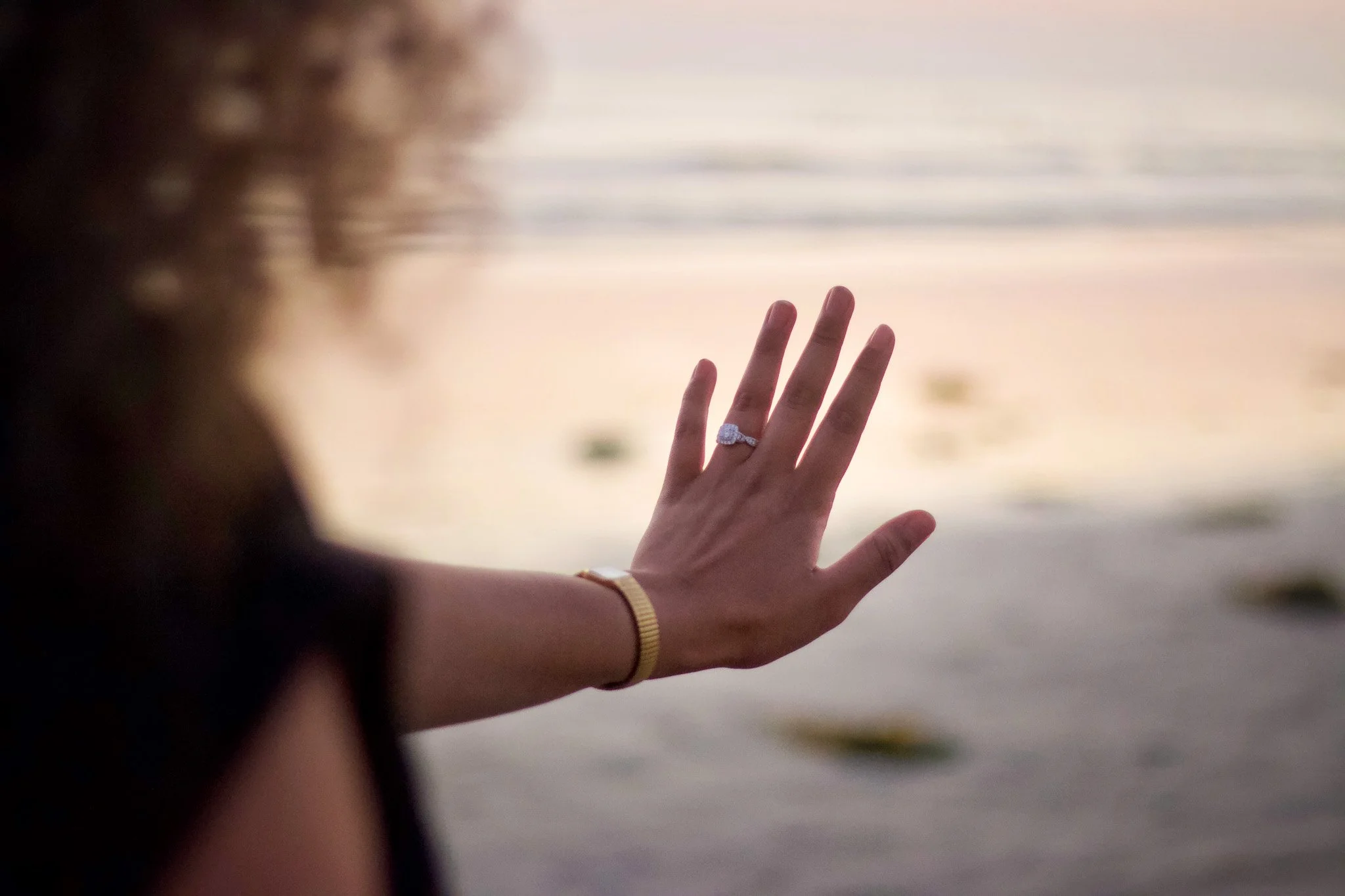 A woman's hand with a diamond ring reaching out towards a body of water during sunset.