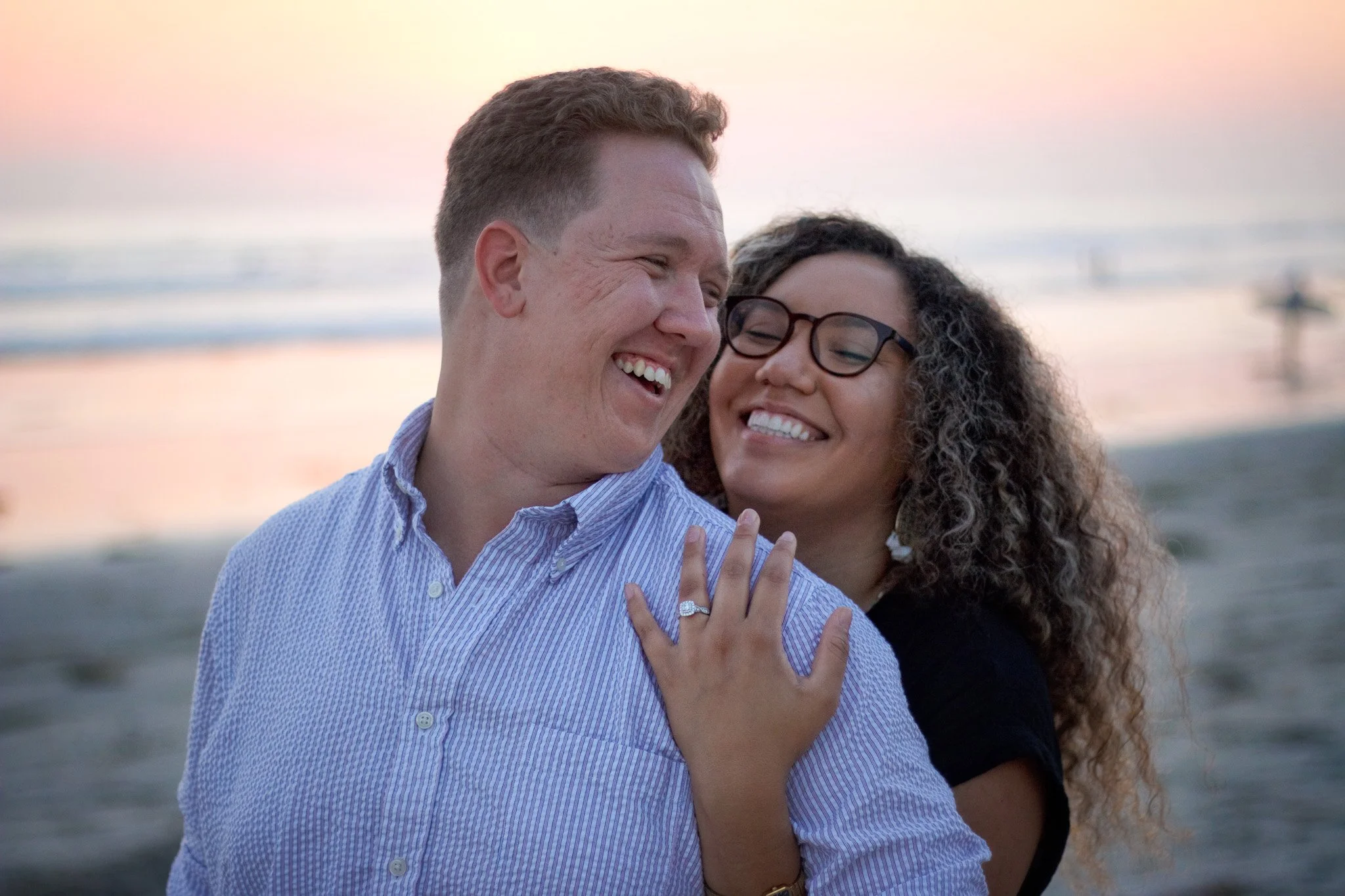 A smiling couple standing on the beach at sunset, with the woman showing her engagement ring on her finger.