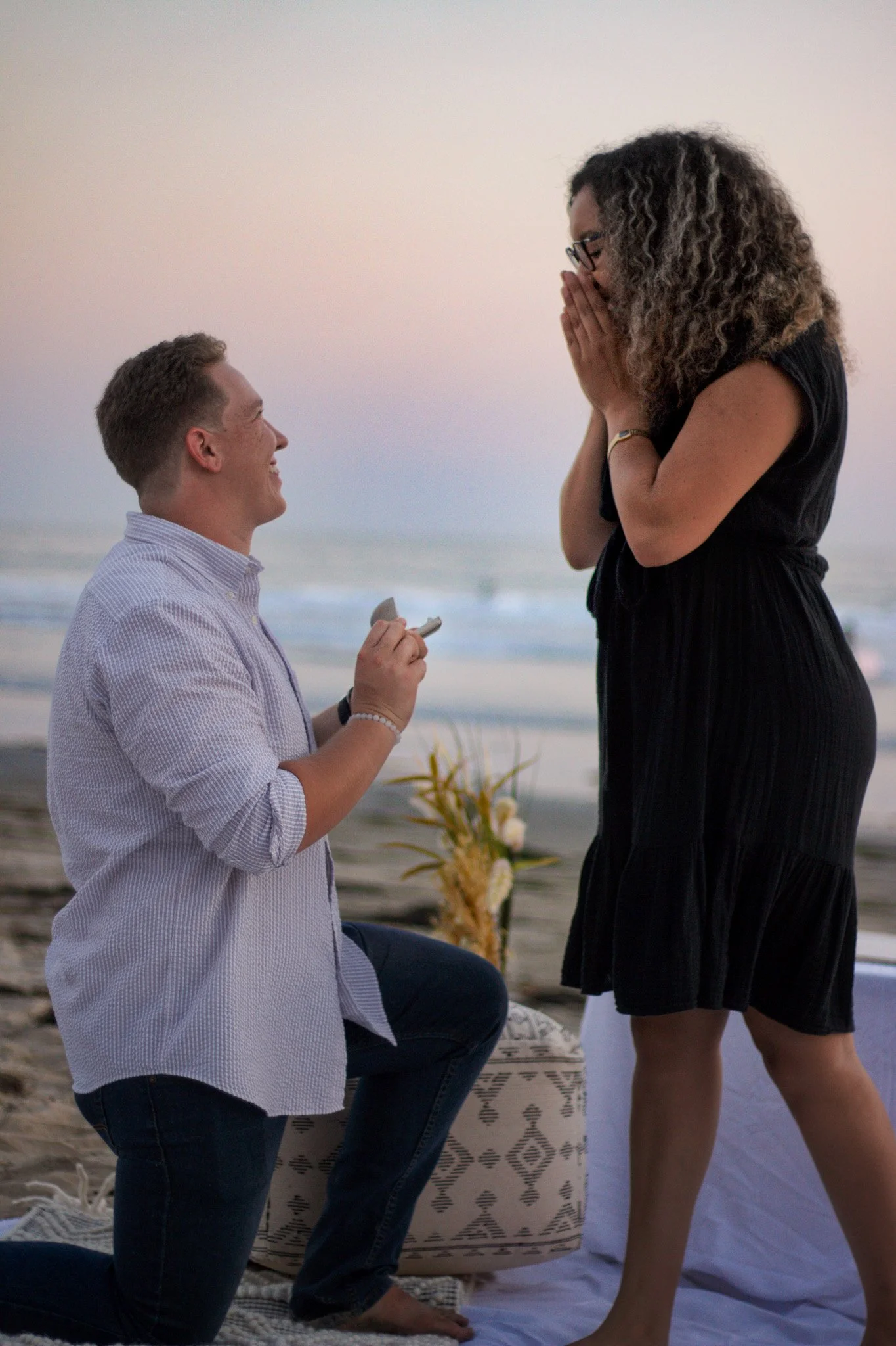 Man proposing marriage to woman on the beach at sunset, woman covers mouth in surprise.