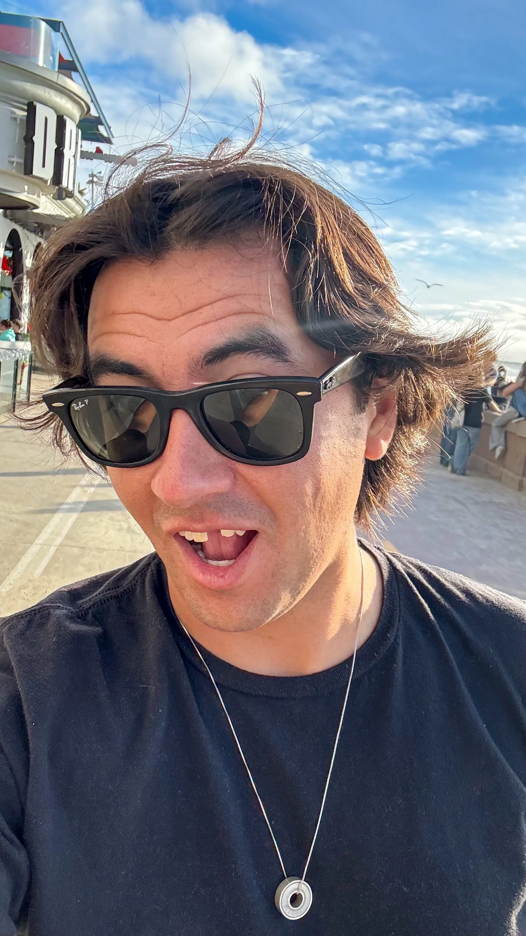 A man wearing sunglasses taking a selfie outdoors on a sunny day, with a blue sky and some clouds behind him. There are people sitting on a concrete ledge and a boat or ferry in the background.