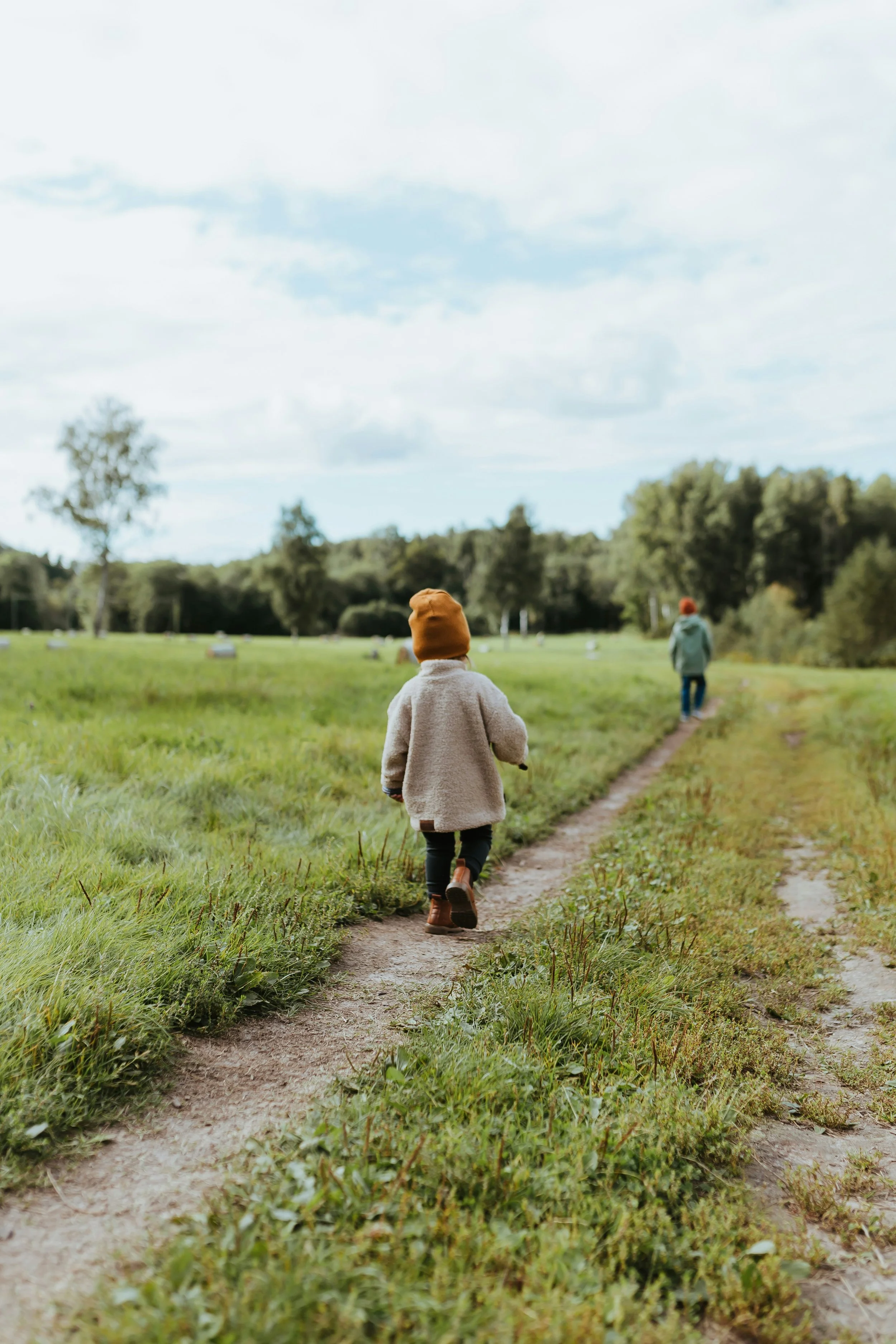 Two children walking on a dirt path through a grassy field with trees and cloudy sky in the background.