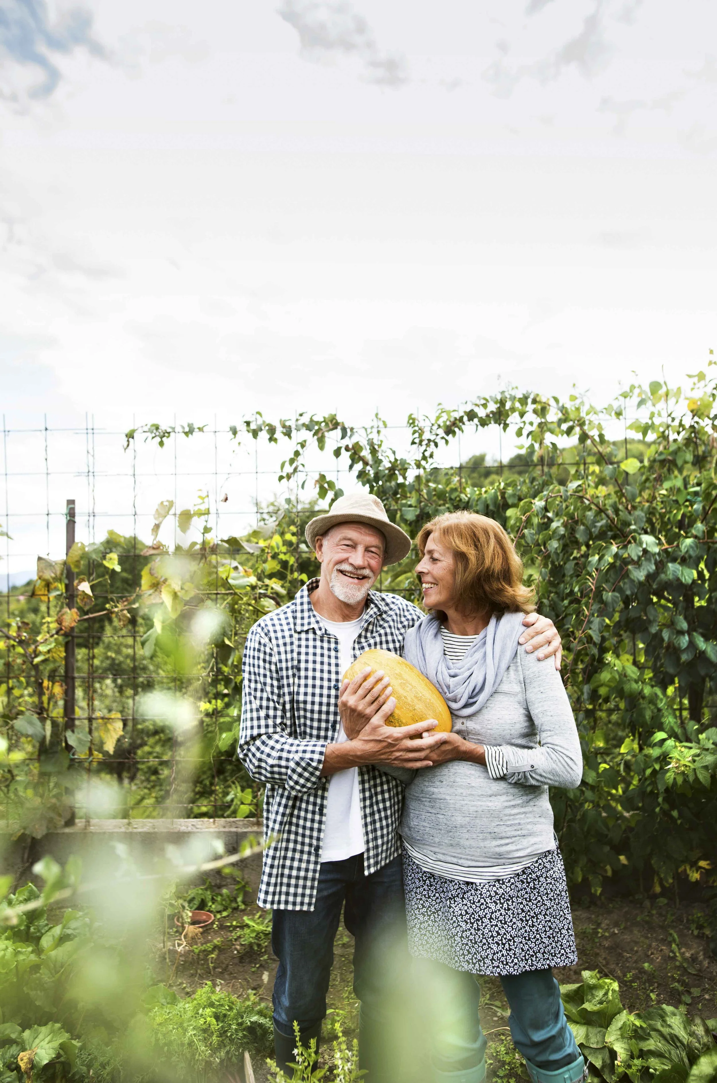 An elderly couple standing in a garden, holding a large yellow pumpkin, smiling at each other.