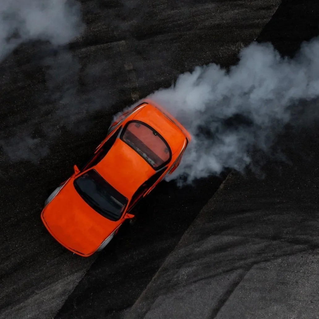 An overhead view of an orange sports car producing white smoke, drifting on a dark asphalt surface.