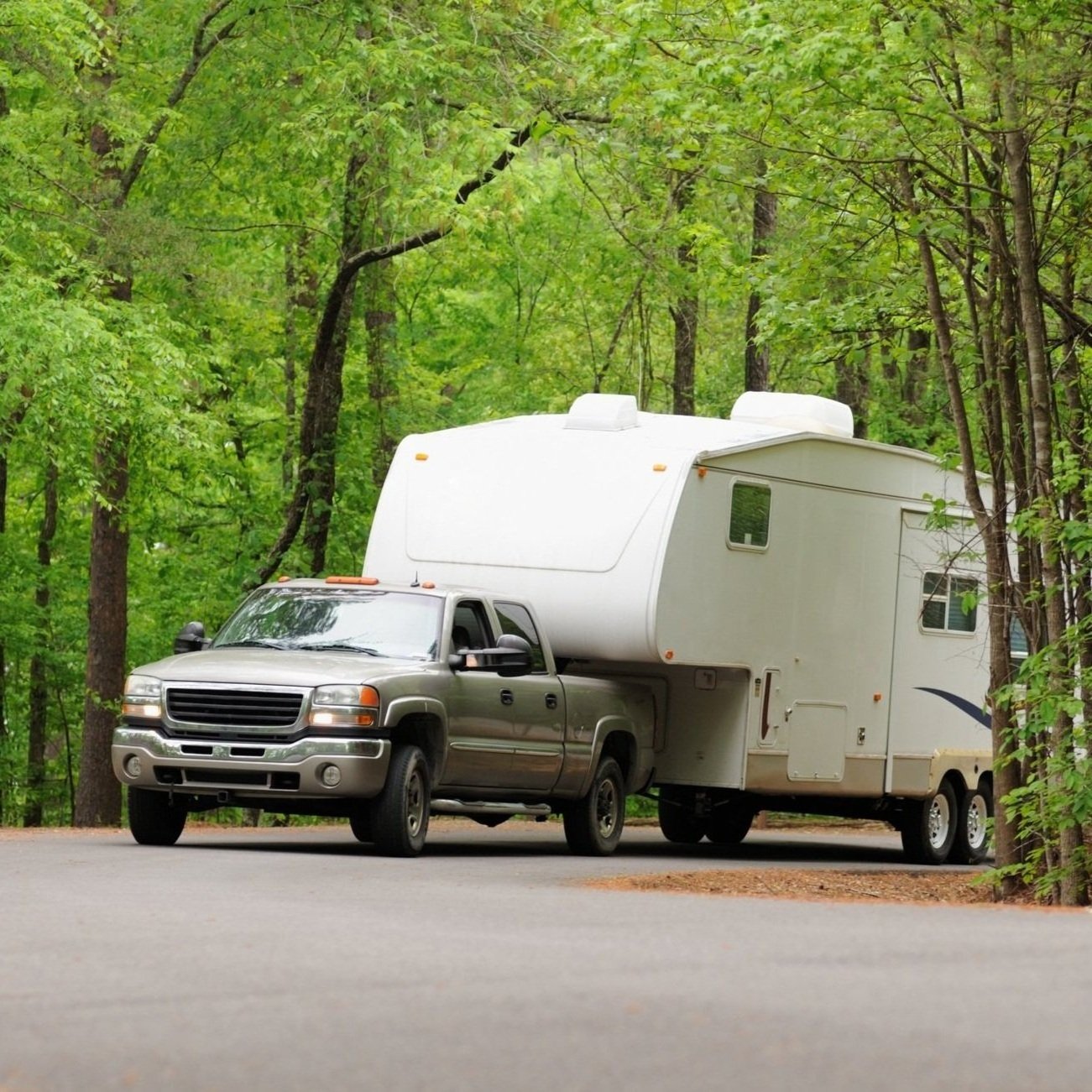 A pickup truck towing a white travel trailer parked on a road surrounded by green trees.