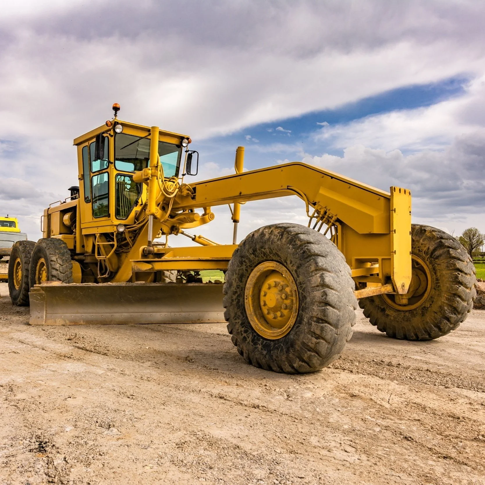 A large yellow construction vehicle, likely a road grader, on dirt ground with cloudy sky in the background.