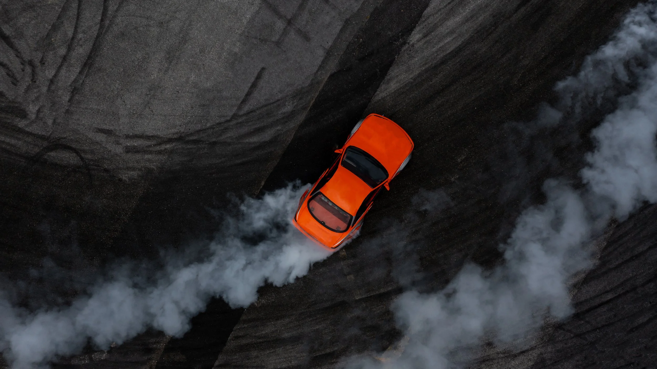 An orange car speeds along a dark, rocky landscape, creating a trail of white smoke behind it.