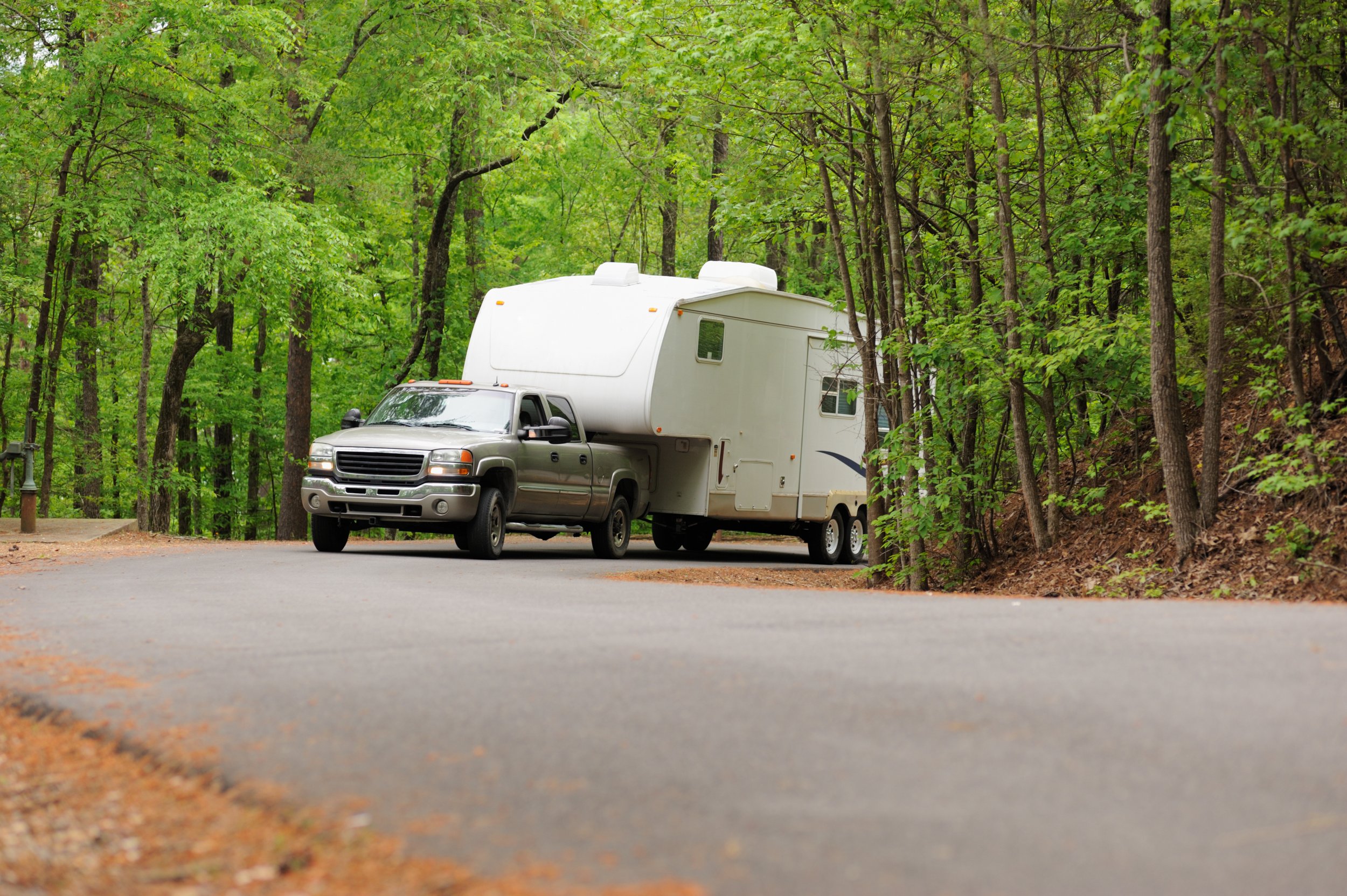 A silver pickup truck towing a large white camper through a forested area with dense green trees.