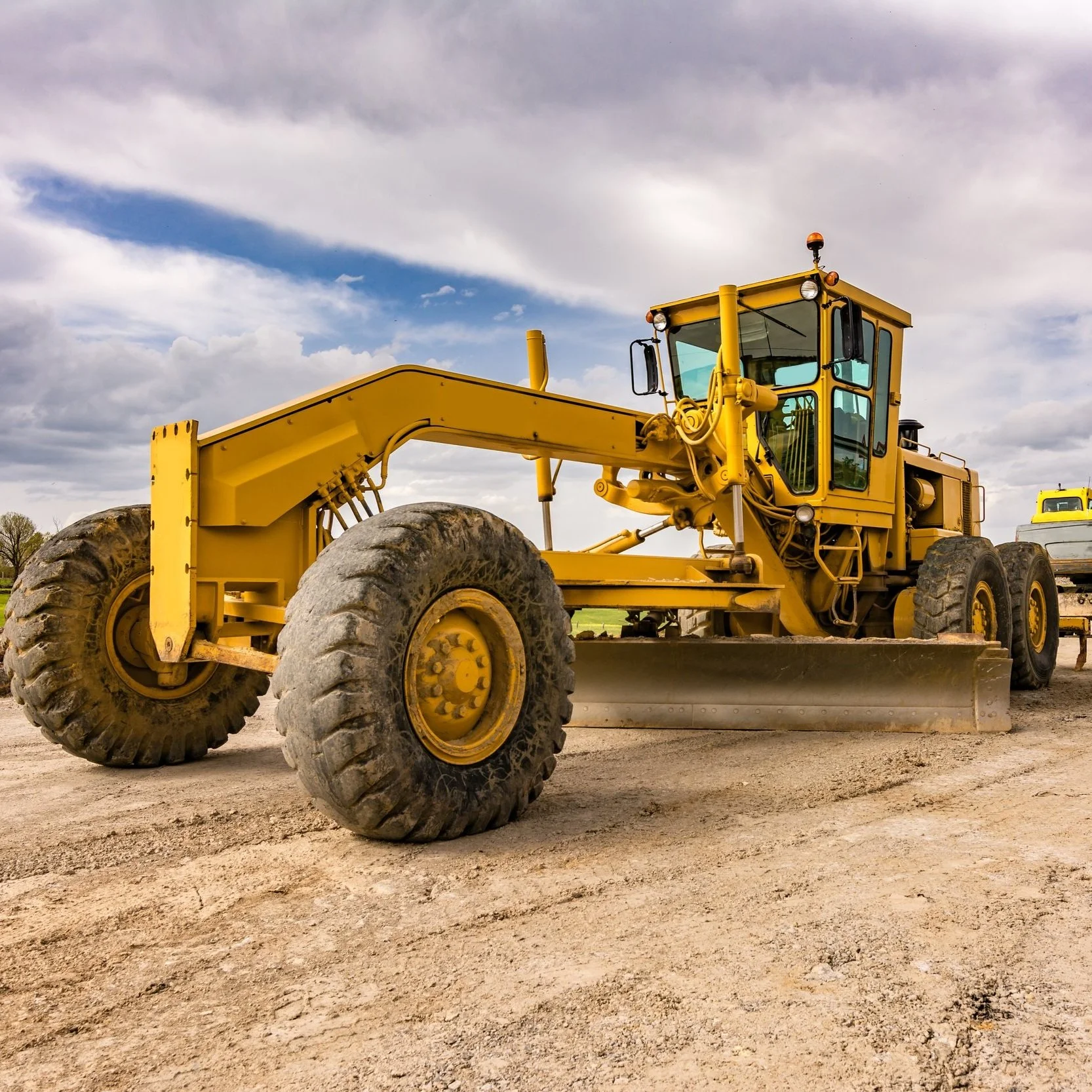 A large yellow construction roller with big dirt-covered tires parked on a dirt lot under a cloudy sky.