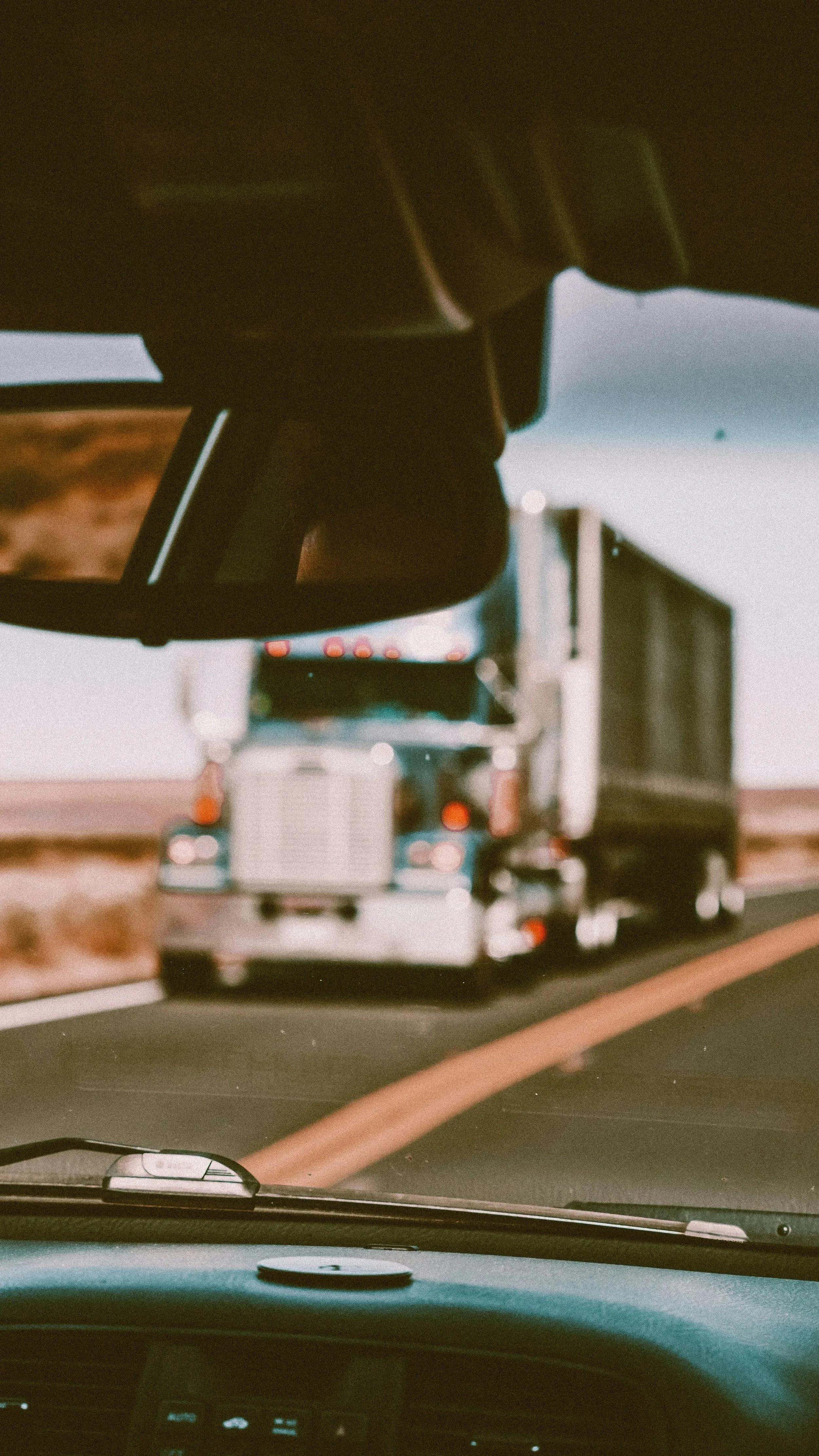 View from inside a vehicle looking at a rearview mirror. The mirror shows a large semi-truck with a white cab and black trailer on the road ahead.