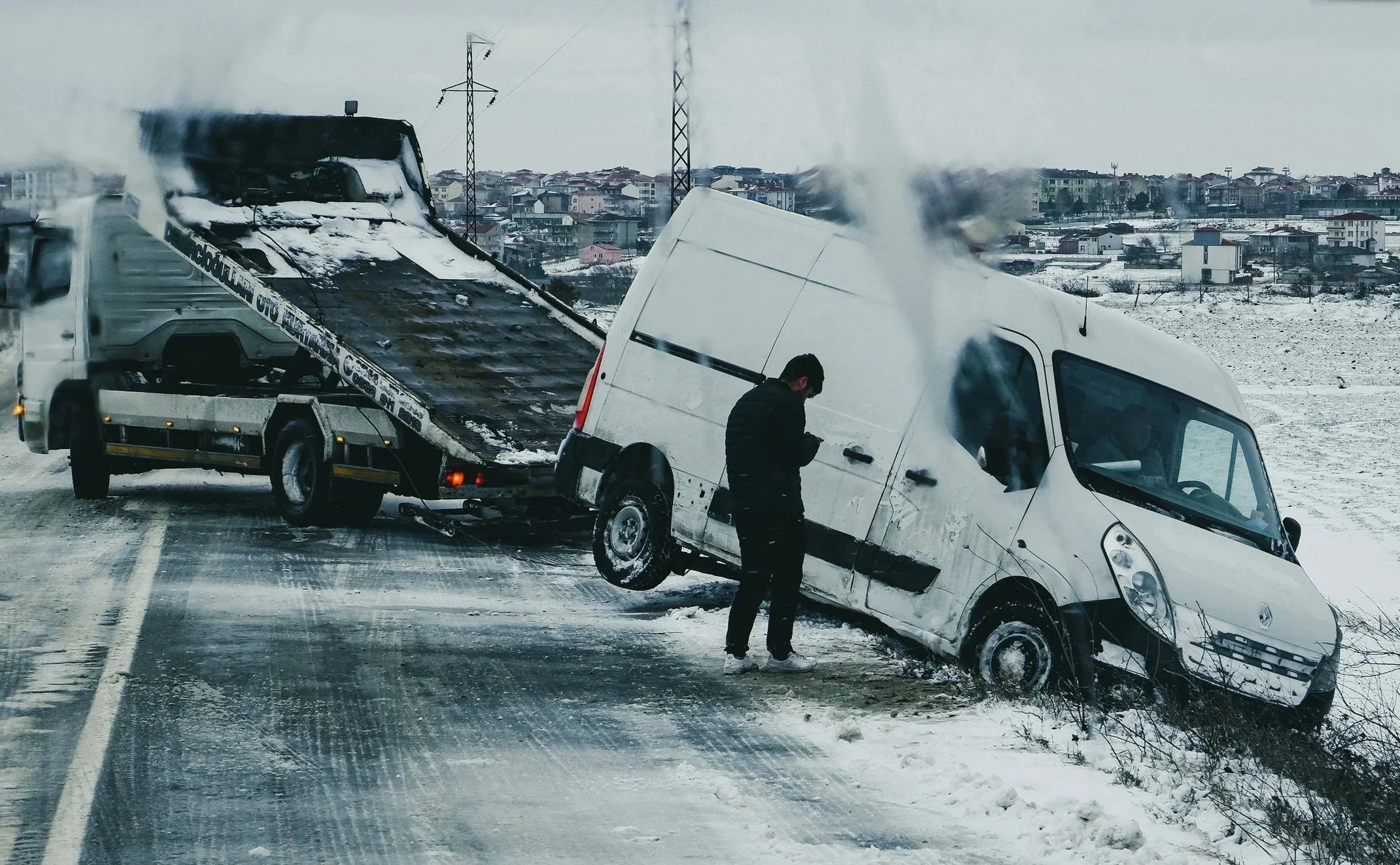 A car accident involving a white van and a black flatbed tow truck on a snowy road. A person is standing next to the van, looking at a phone, with smoke or steam rising from the vehicles. Snow-covered houses and power lines are visible in the background.