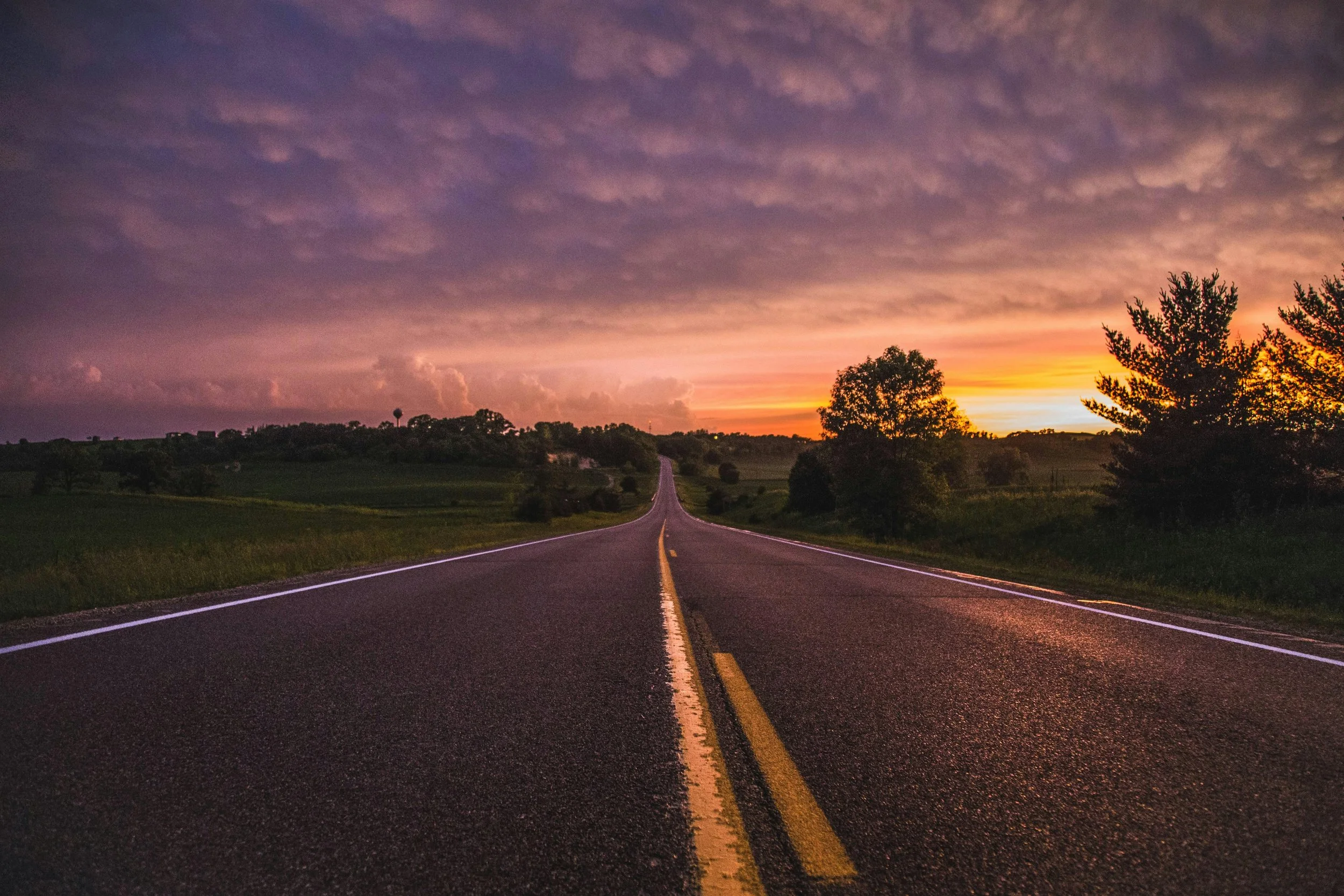 A two-lane road stretching into the distance during sunset, with trees on both sides and colorful clouds in the sky.