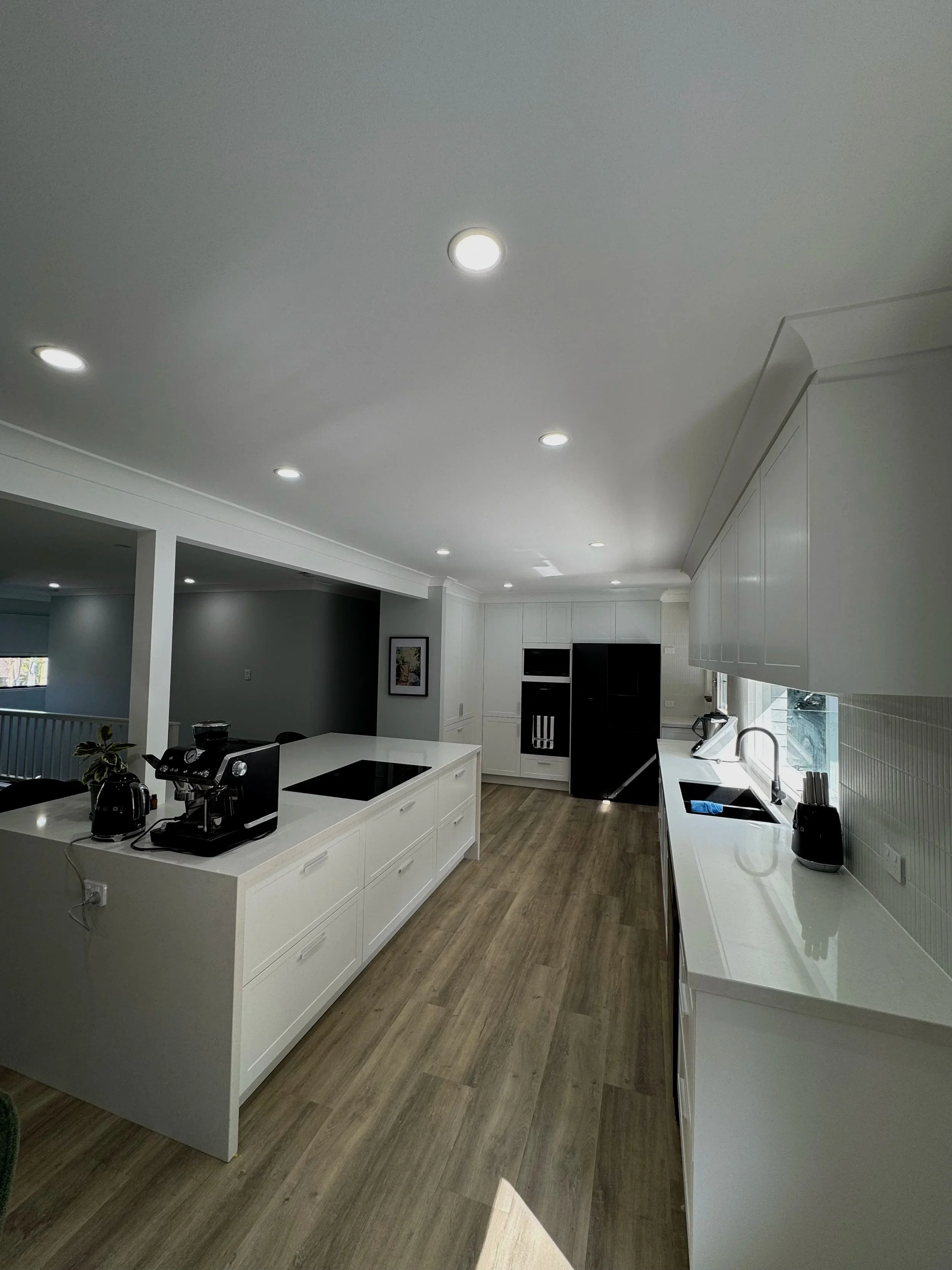 Modern kitchen with white cabinets, a black refrigerator, an island with a cooktop, and a sink under a window. Wooden flooring and recessed ceiling lights.