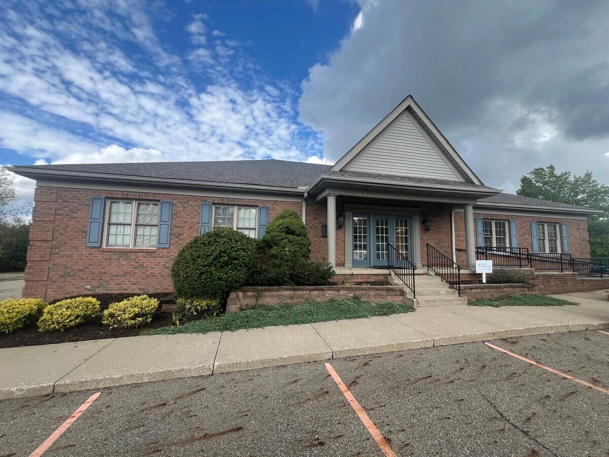 Brick building with a gabled roof, front steps, and three sets of double doors, with parking spaces and cloudy sky.