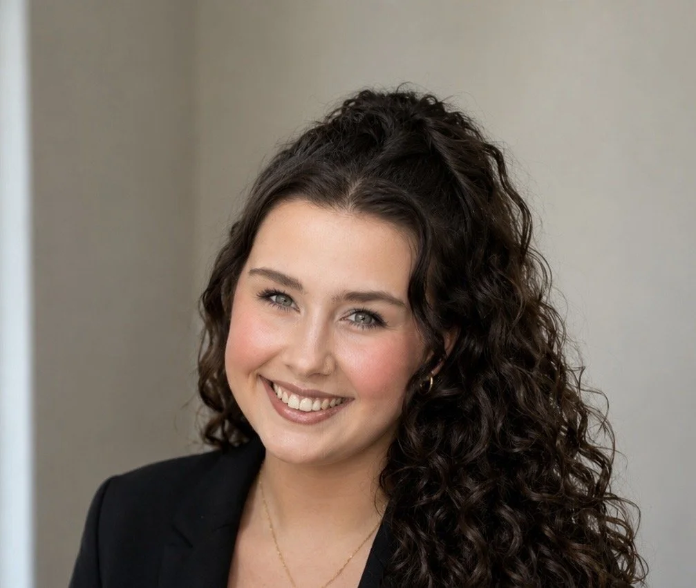 A smiling woman with long, curly dark hair, wearing a black blazer and gold jewelry, posed against a neutral background.