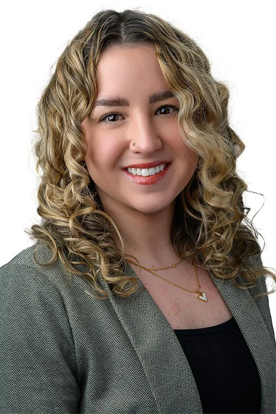 A smiling woman with curly blonde hair, wearing a gray blazer and a black top, accessorized with a gold necklace and a nose piercing.