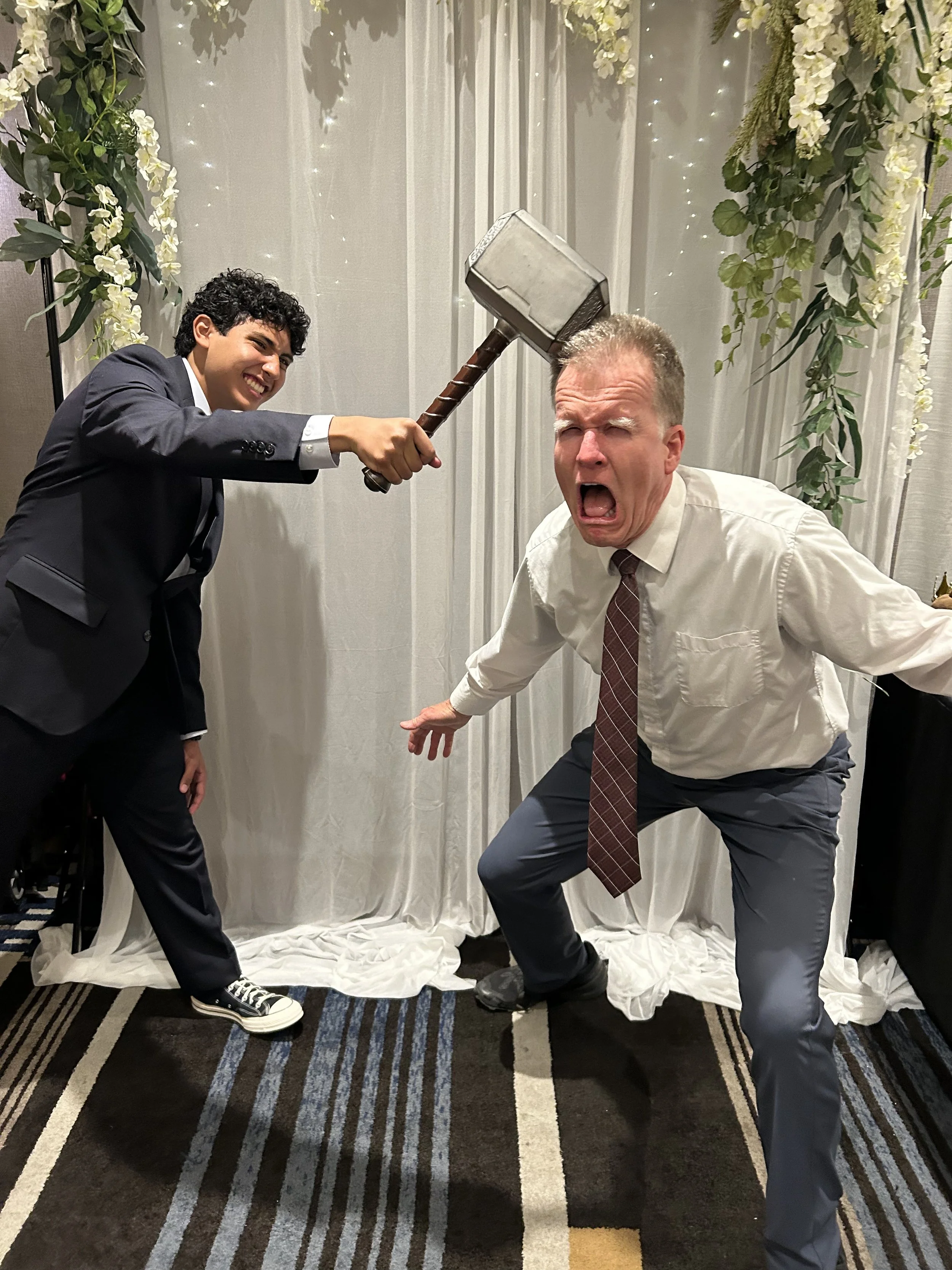 Two men engaged in playful mock fight, one with a hammer and the other with a surprised expression, in front of a white backdrop decorated with floral curtains.