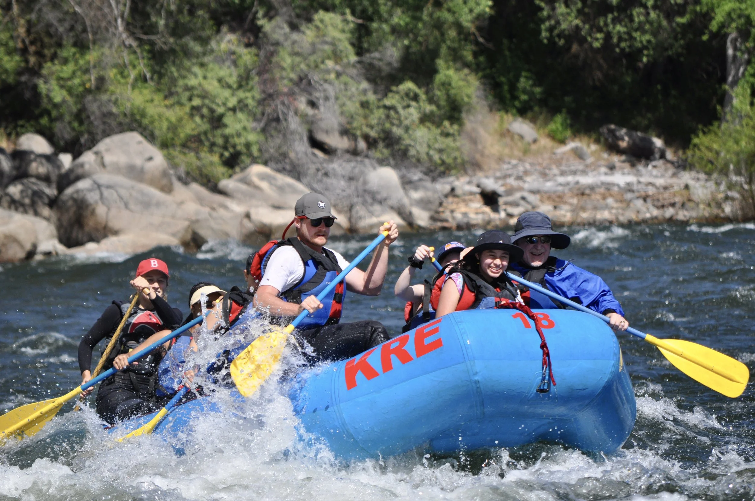 Group of people white-water rafting on a river with rocks and green trees in the background, wearing life jackets and holding paddles.