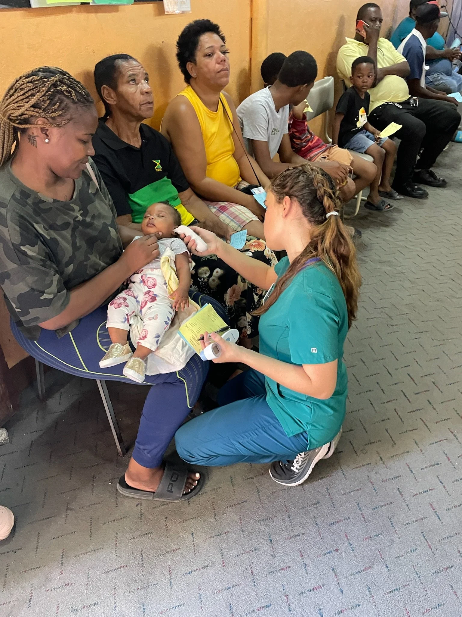 A group of people sitting on chairs against a yellow wall, with a healthcare worker checking the pulse of a baby lying on her lap. The baby is dressed in floral pajamas and appears to be resting. The group includes children and adults, some holding p