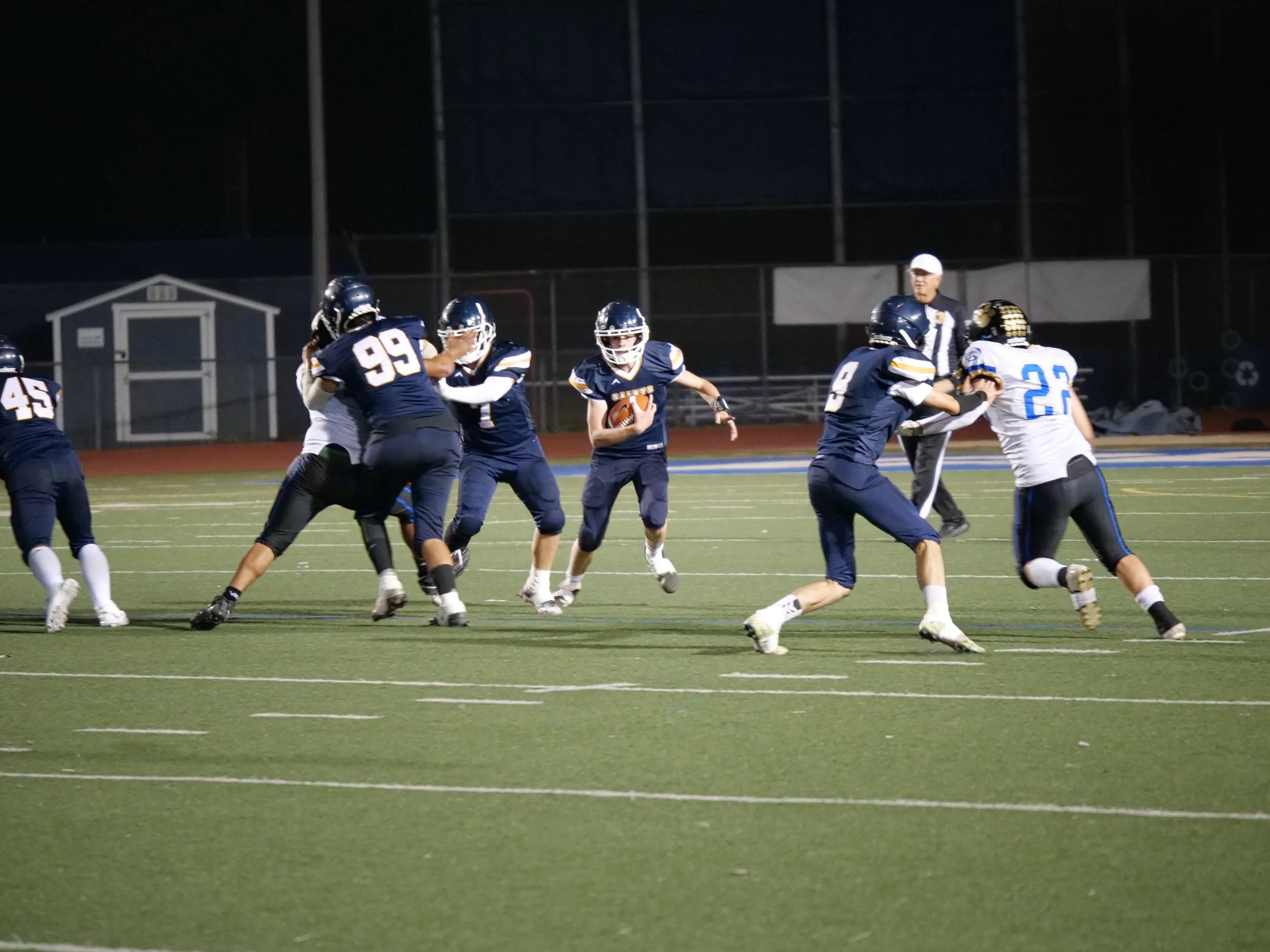A nighttime American football game showing players in navy blue and white uniforms, with the team name 'Cougars' on their jerseys, engaged in a play on the field while a referee observes.