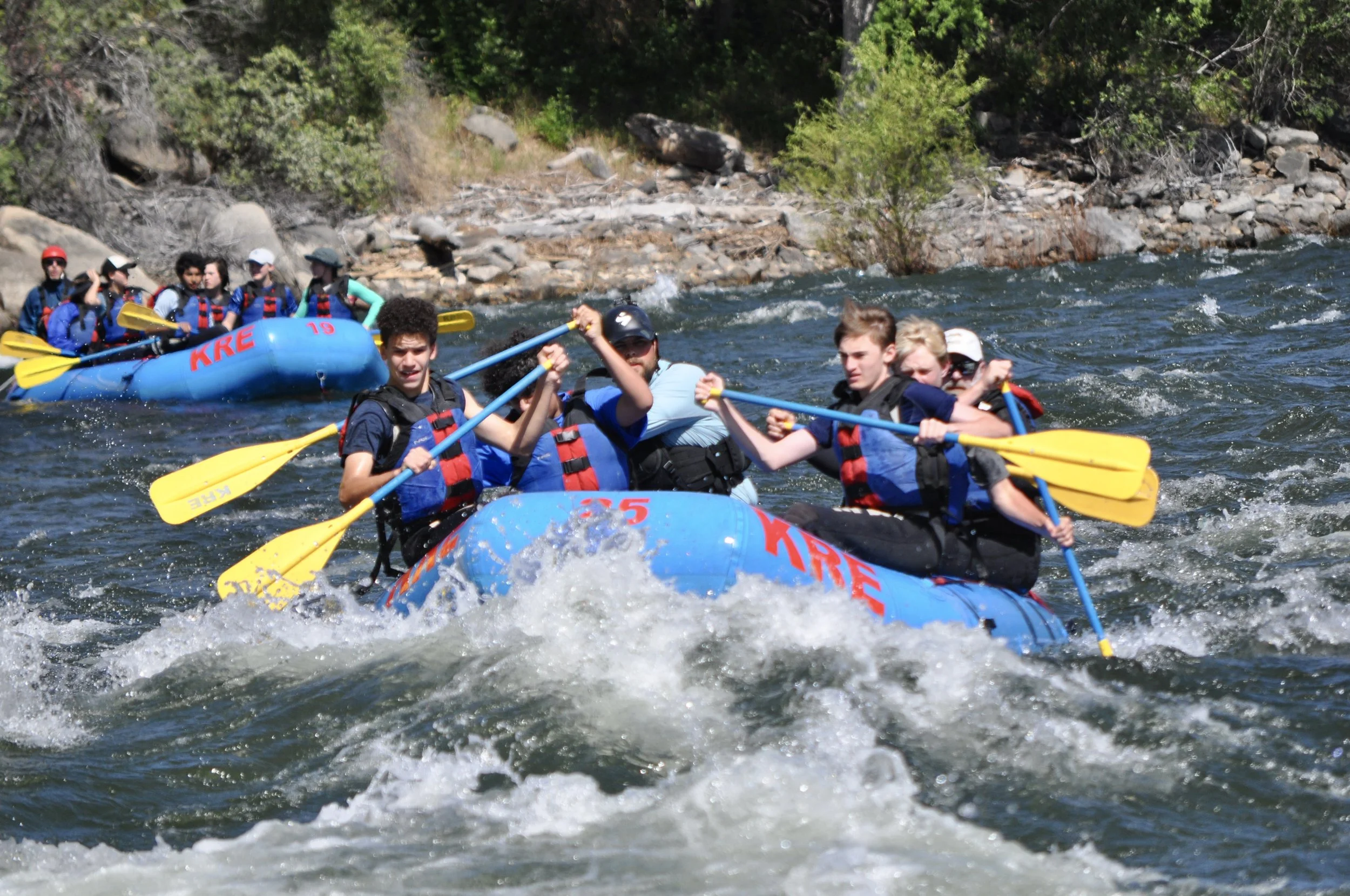 A group of people rafting on a river with rocks and greenery in the background.