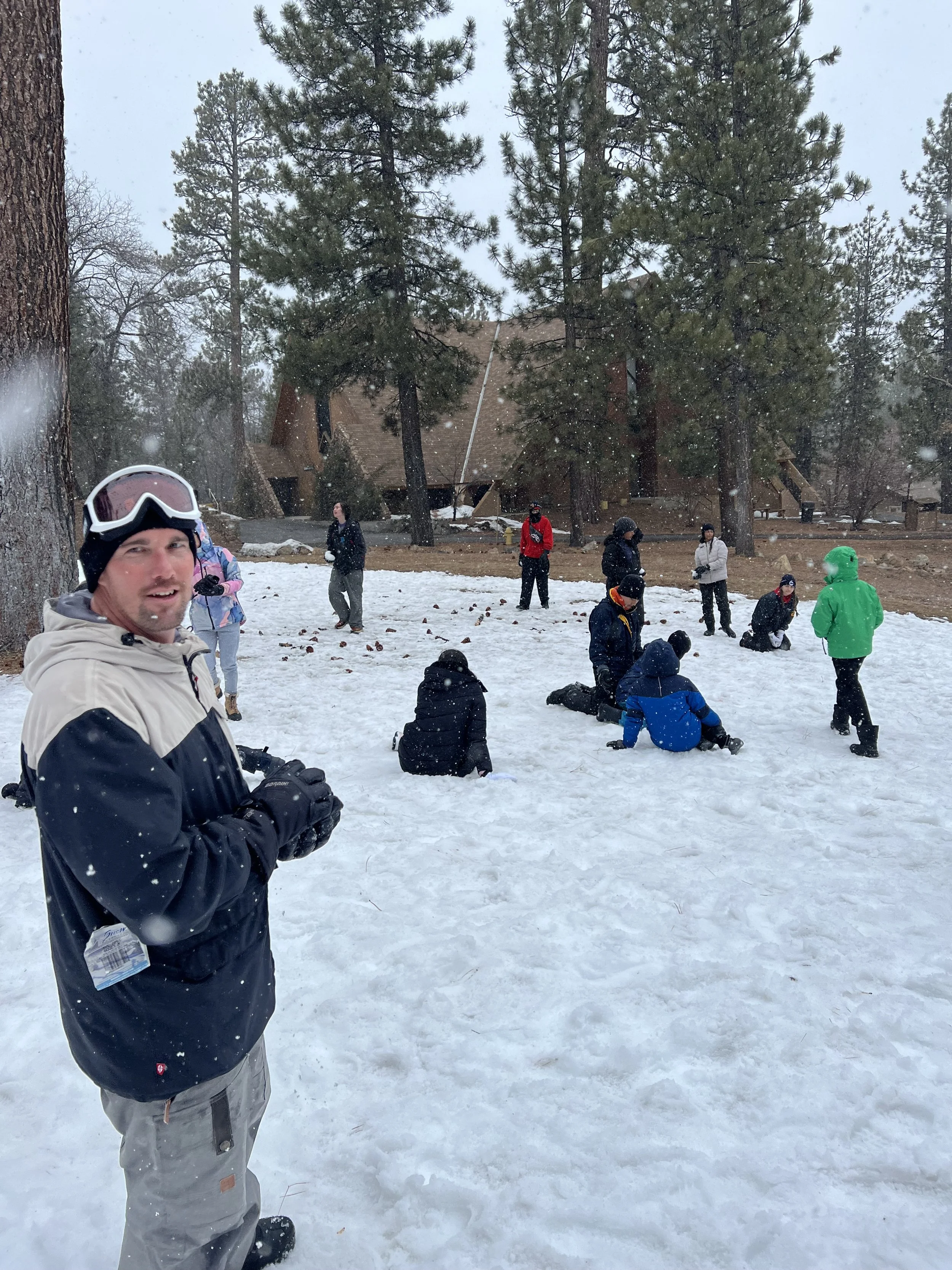 A group of people gathered in a snowy forested area, some sitting or kneeling on the snow, others standing, with trees and a house in the background. A man in the foreground is smiling and wearing a ski jacket and goggles.