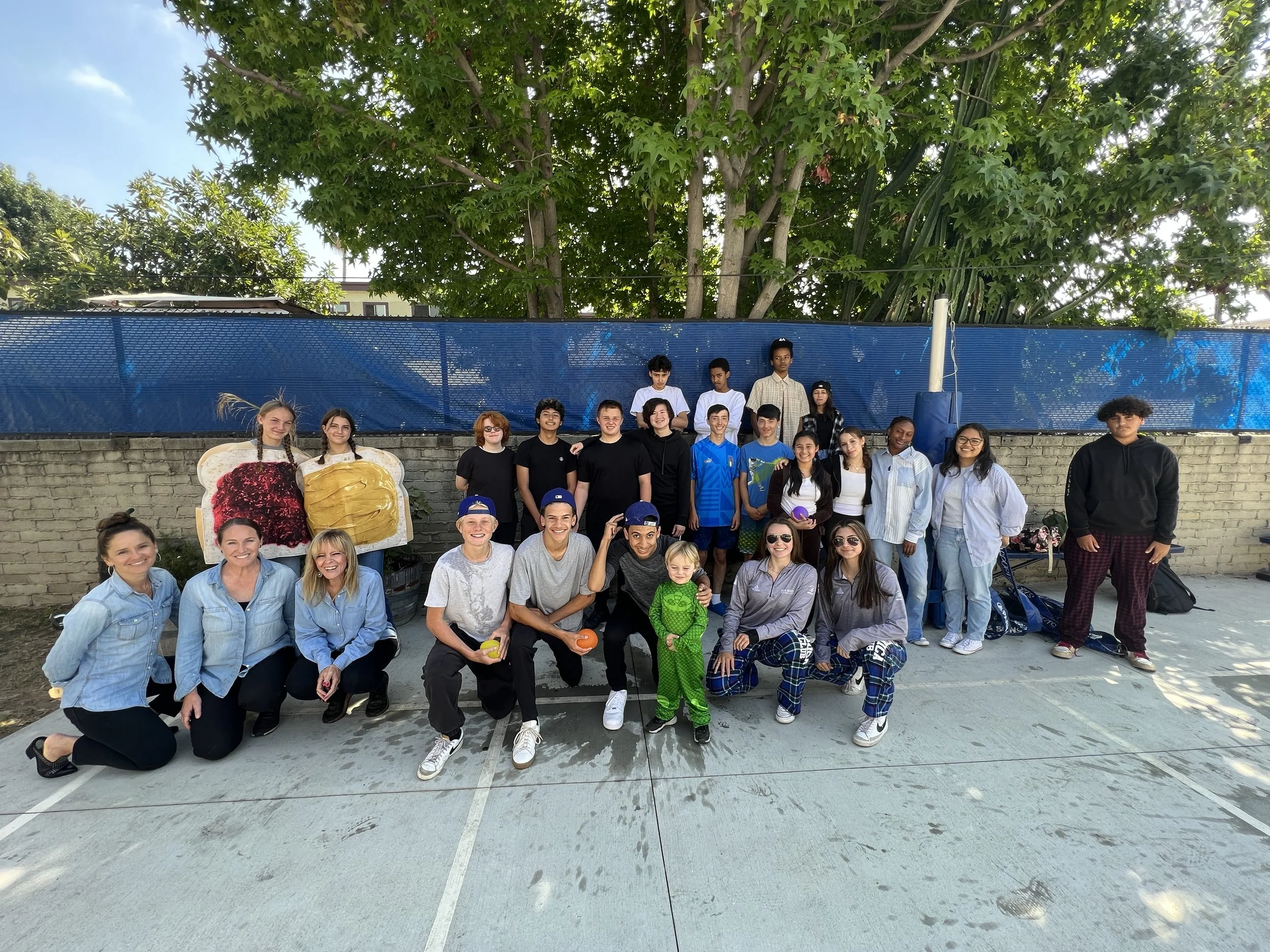 Group of diverse children and adults standing together outdoors in front of a blue fence, with large green trees and a building in the background, enjoying a sunny day.