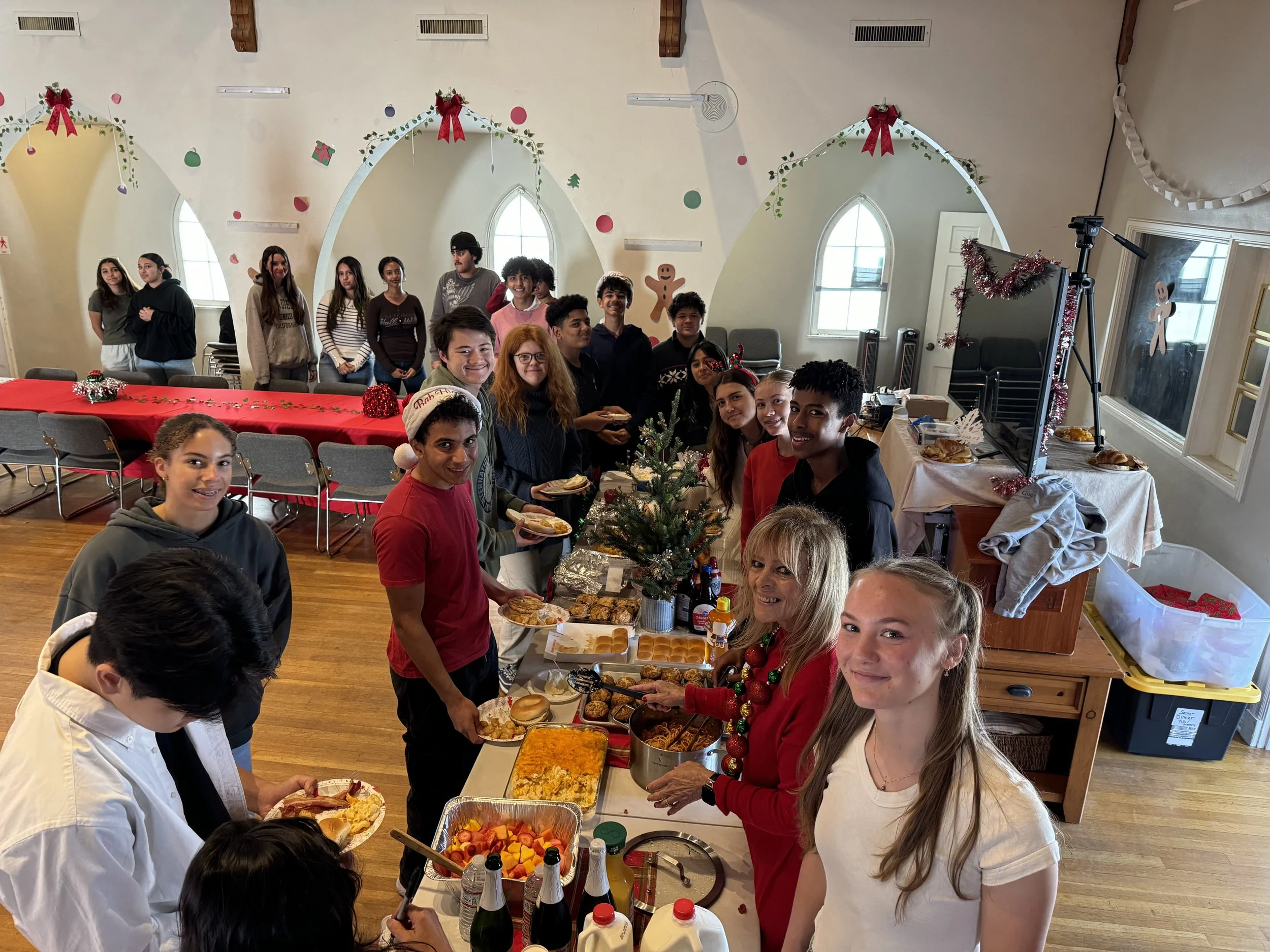 Group of children and teenagers gathered around a table with food and drinks, celebrating Christmas in a decorated room with holiday ornaments and a small Christmas tree.