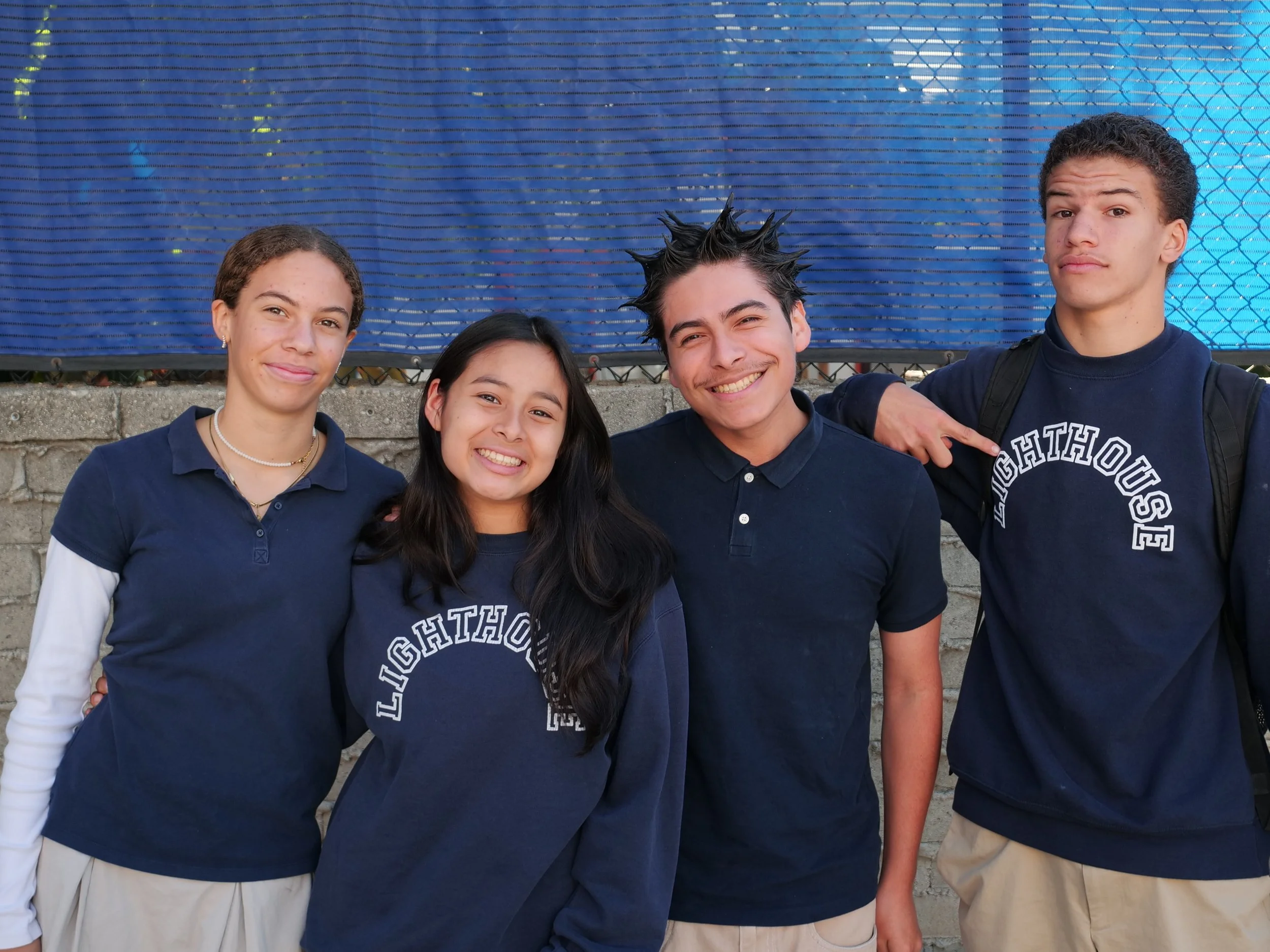 Four teenagers standing outdoors in front of a blue fence, smiling at the camera. Three of them are girls and one is a boy. They are wearing navy blue and white clothing.