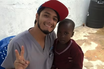 A young man in medical scrubs and a cap making a victory sign with his hand, standing next to a young girl with a serious expression, outdoors with a white wall and trash bin in the background.