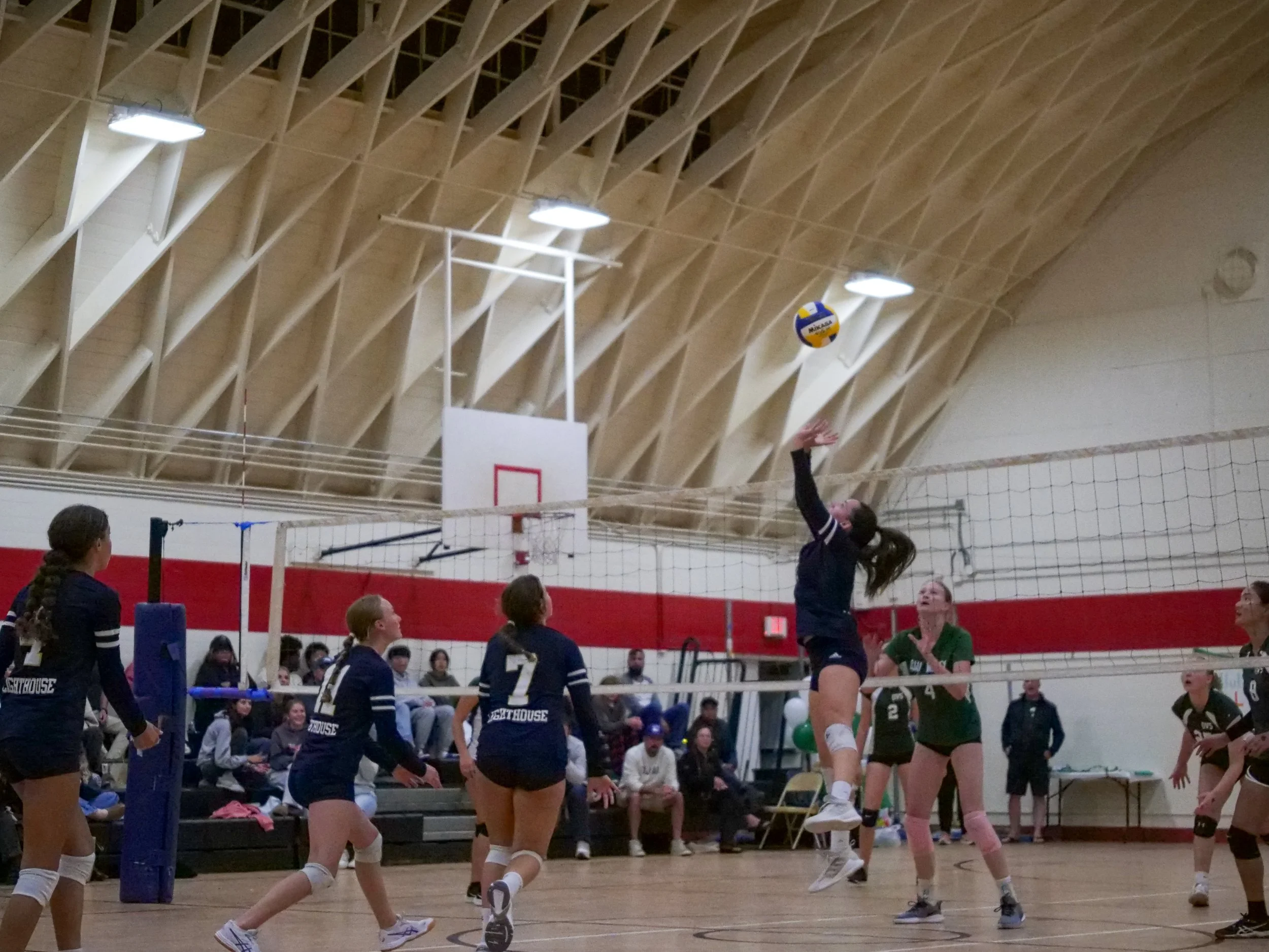 Indoor volleyball game with players on both sides, some jumping to hit the ball over the net, spectators watching from the benches and seating area.