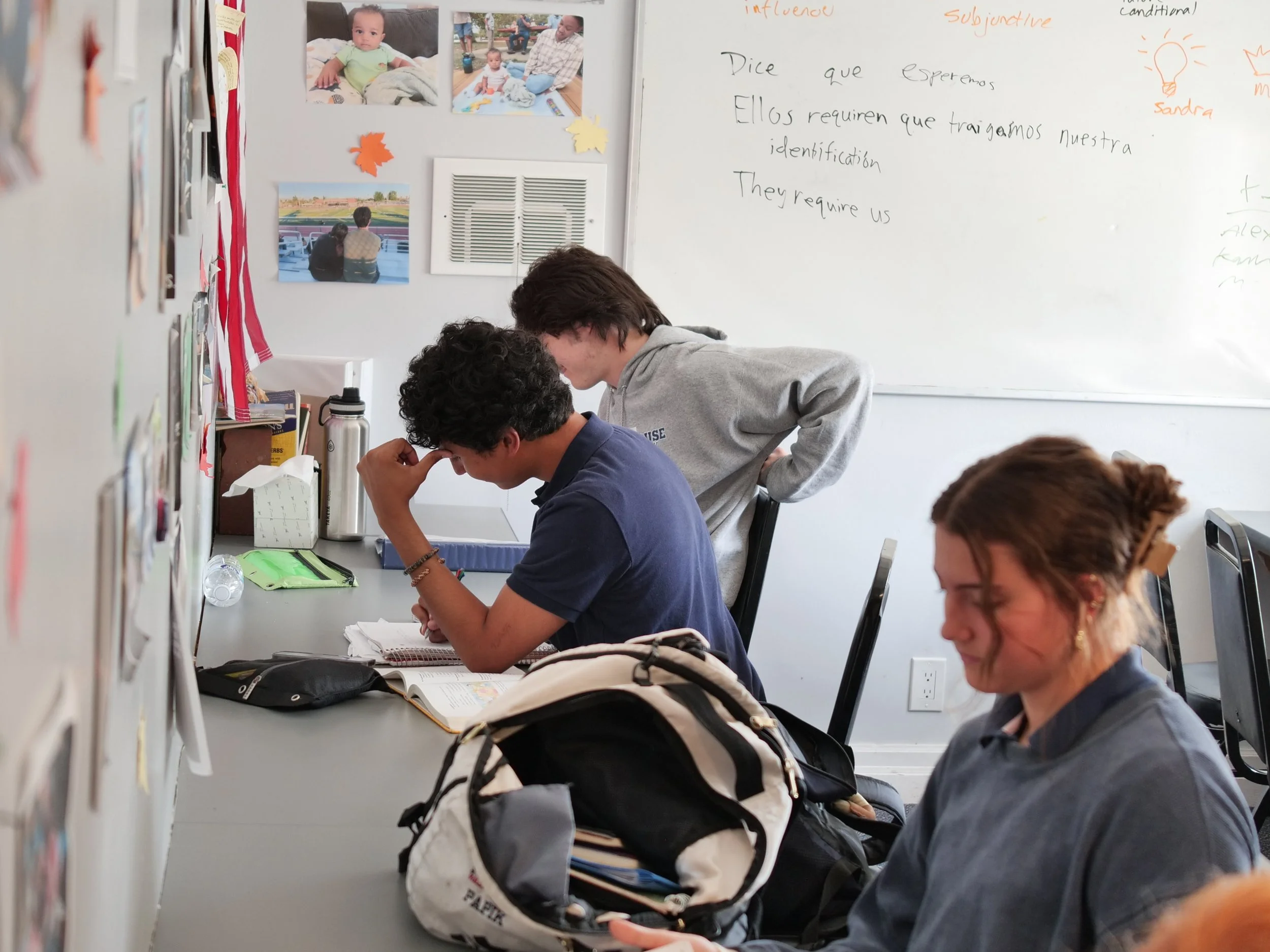 Students seated at desks in a classroom, engaged in a learning activity, with backpacks on the table and photos on the wall.
