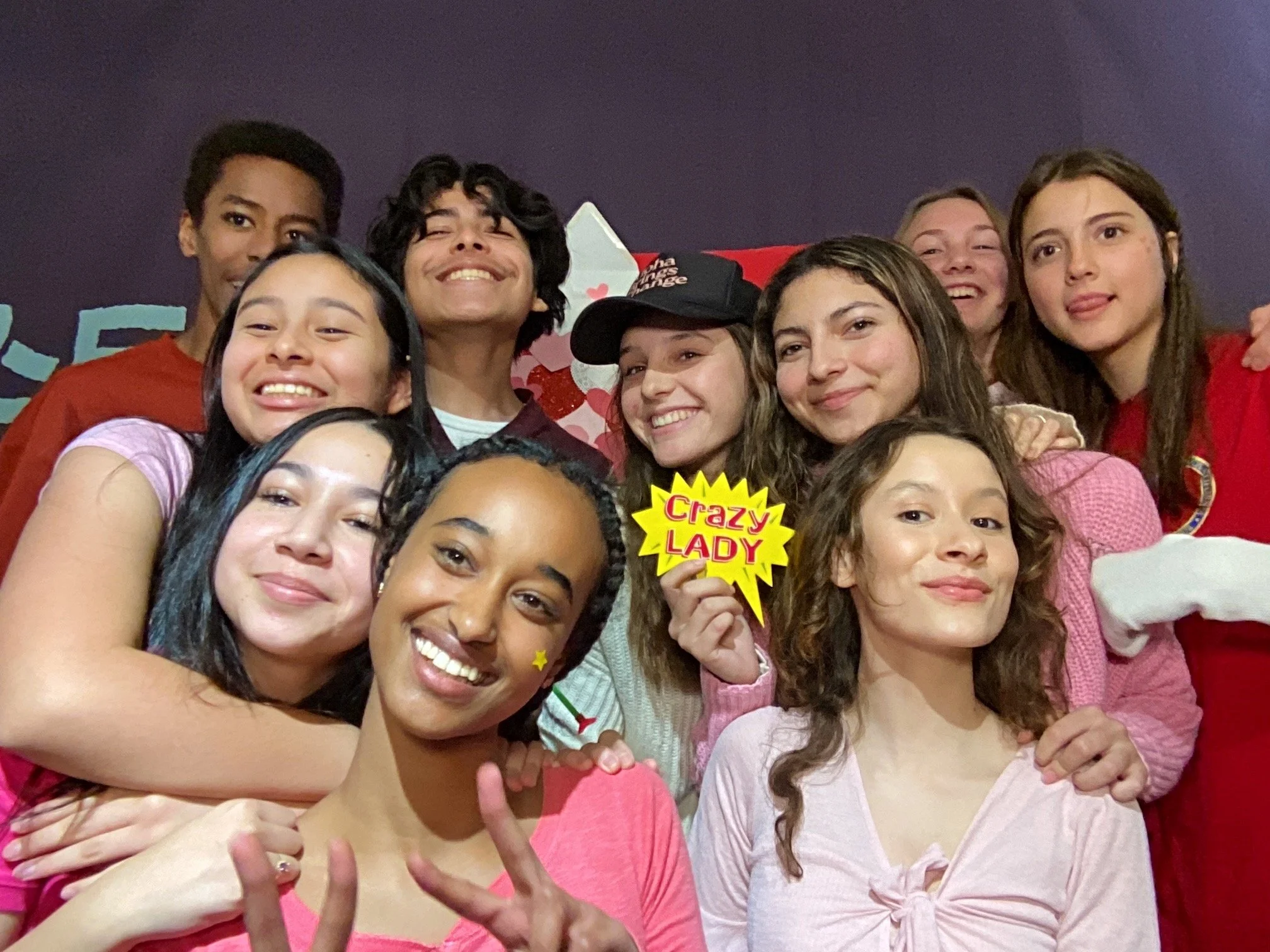 Group of smiling young women and men posing closely for a selfie, some holding colorful speech bubble props with words like 'Crazy Lady'. The background is purple and red with some holiday-themed decorations.