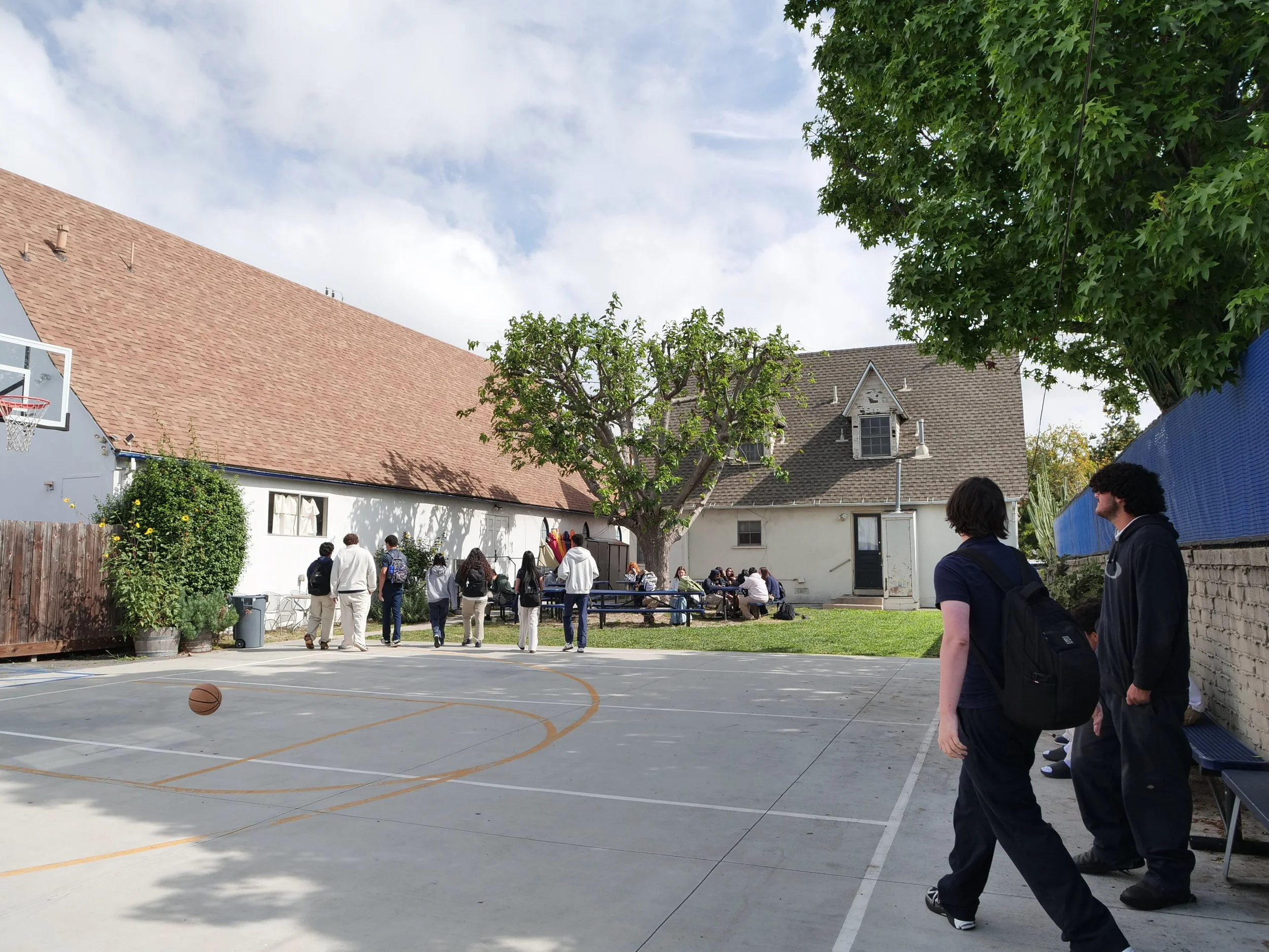 Schoolyard with students and teachers gathered, some sitting at picnic tables, others standing on a basketball court. The scene includes a large tree, a basketball hoop, a white building, and a house in the background under a partly cloudy sky.
