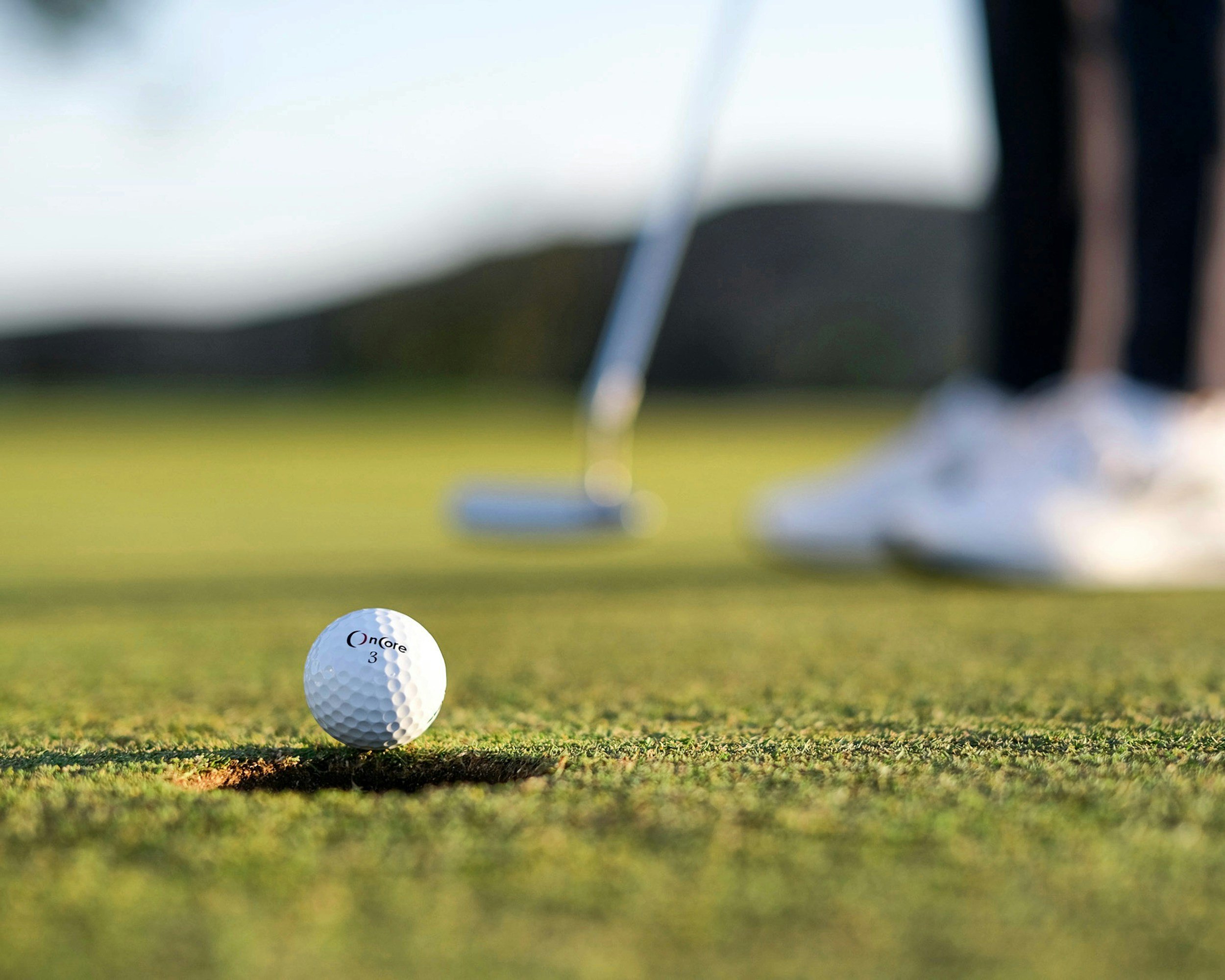 Close-up of a golf ball on the green grass at a golf course, with a blurred background including a golfer's legs and a golf club.
