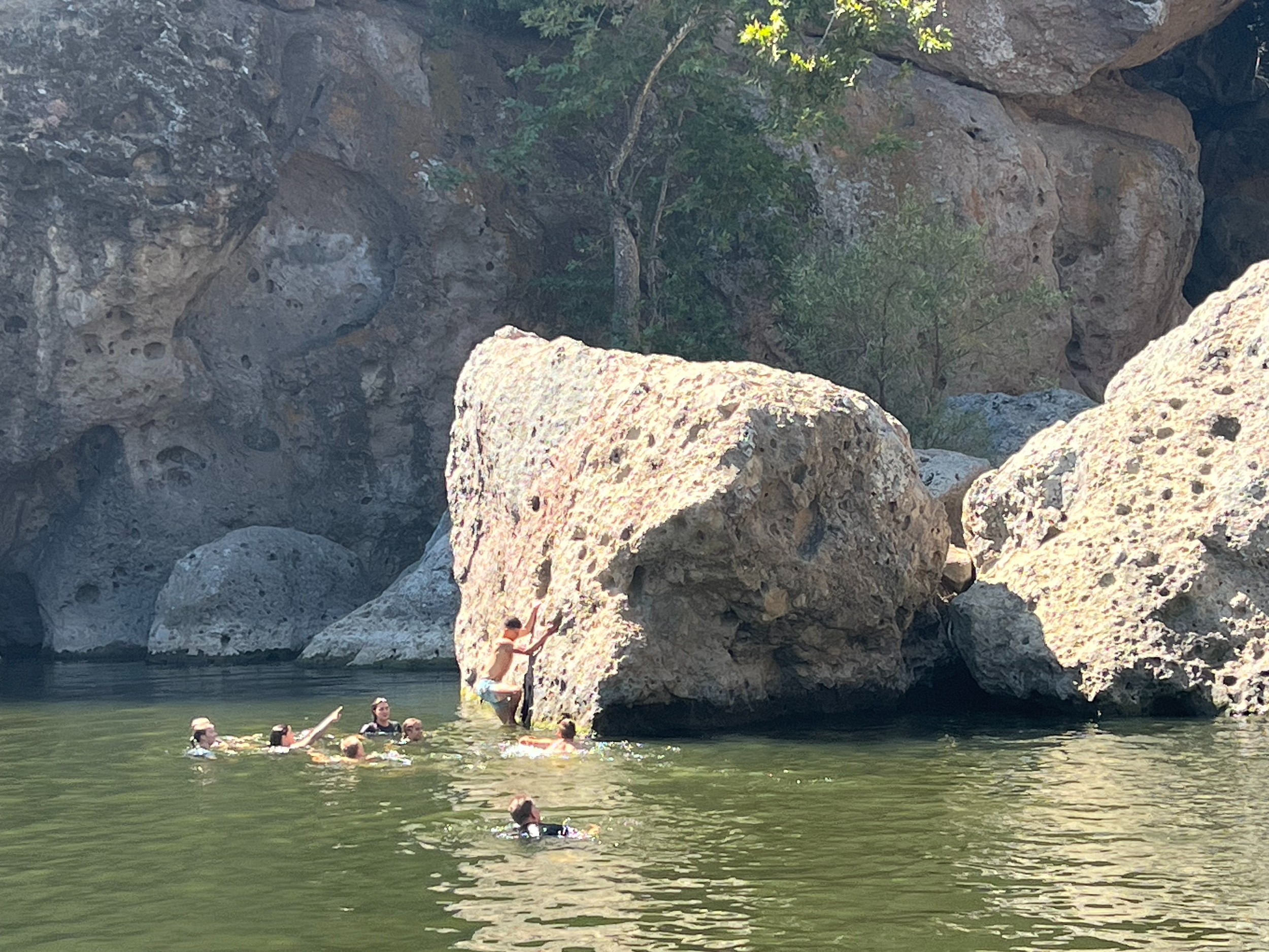 People swimming and relaxing in a river at the base of rocky cliffs with trees and sunlight overhead.