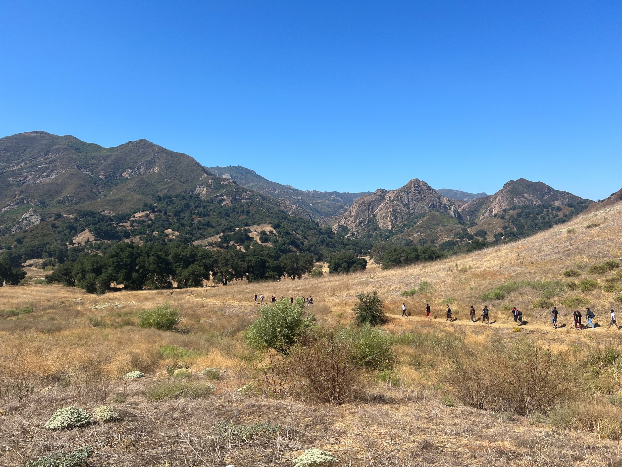 A group of people walking in a line through a dry, grassy field with mountains in the background under a clear blue sky.