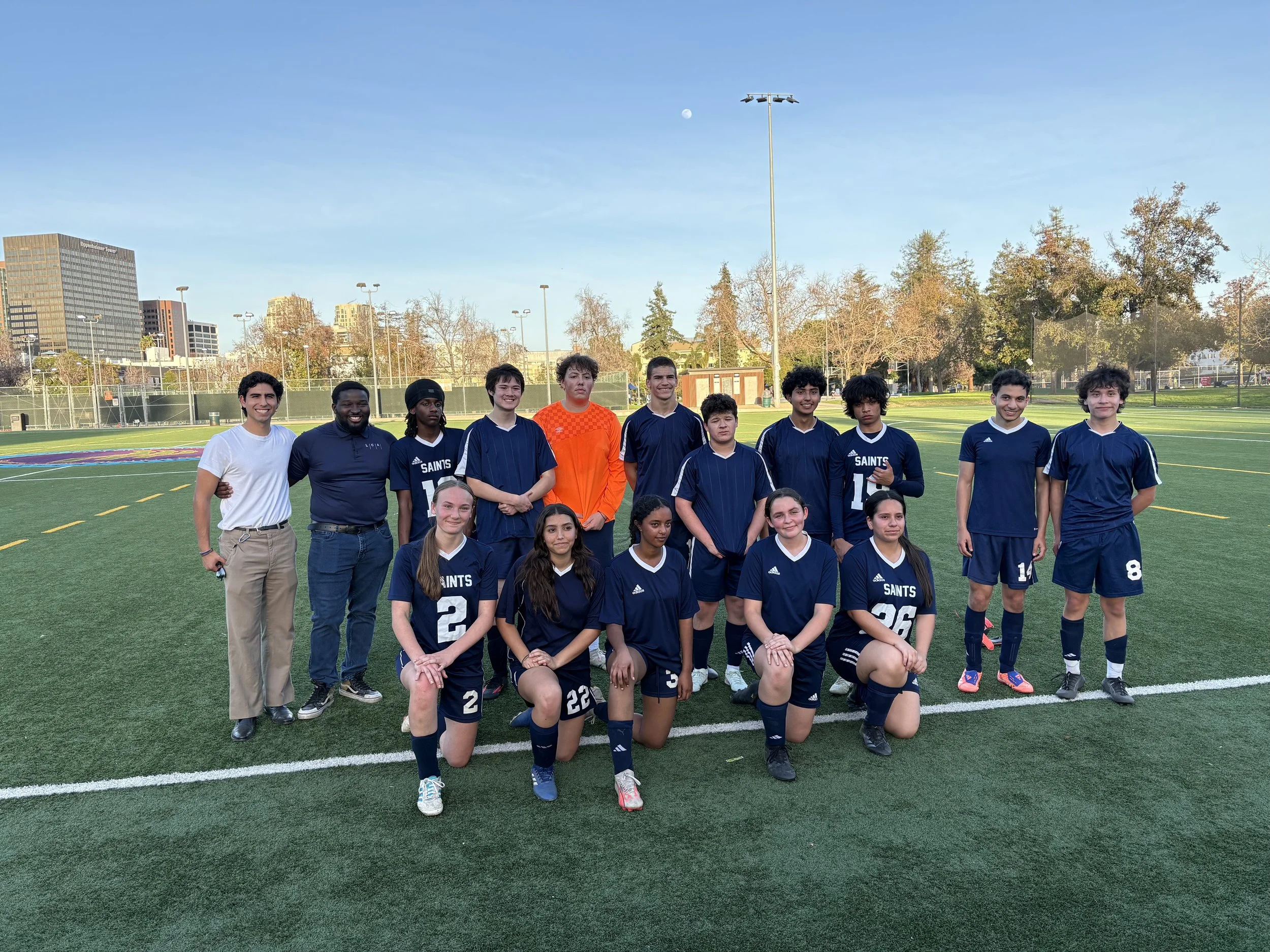 A group of young soccer players and their coaches standing on a soccer field in a team photo. The players are wearing navy blue uniforms with white accents, and some are kneeling in the front row while others stand behind. The coaches stand to the left and right sides of the group. The background features a green field, a fence, tall trees, and buildings under a clear blue sky.