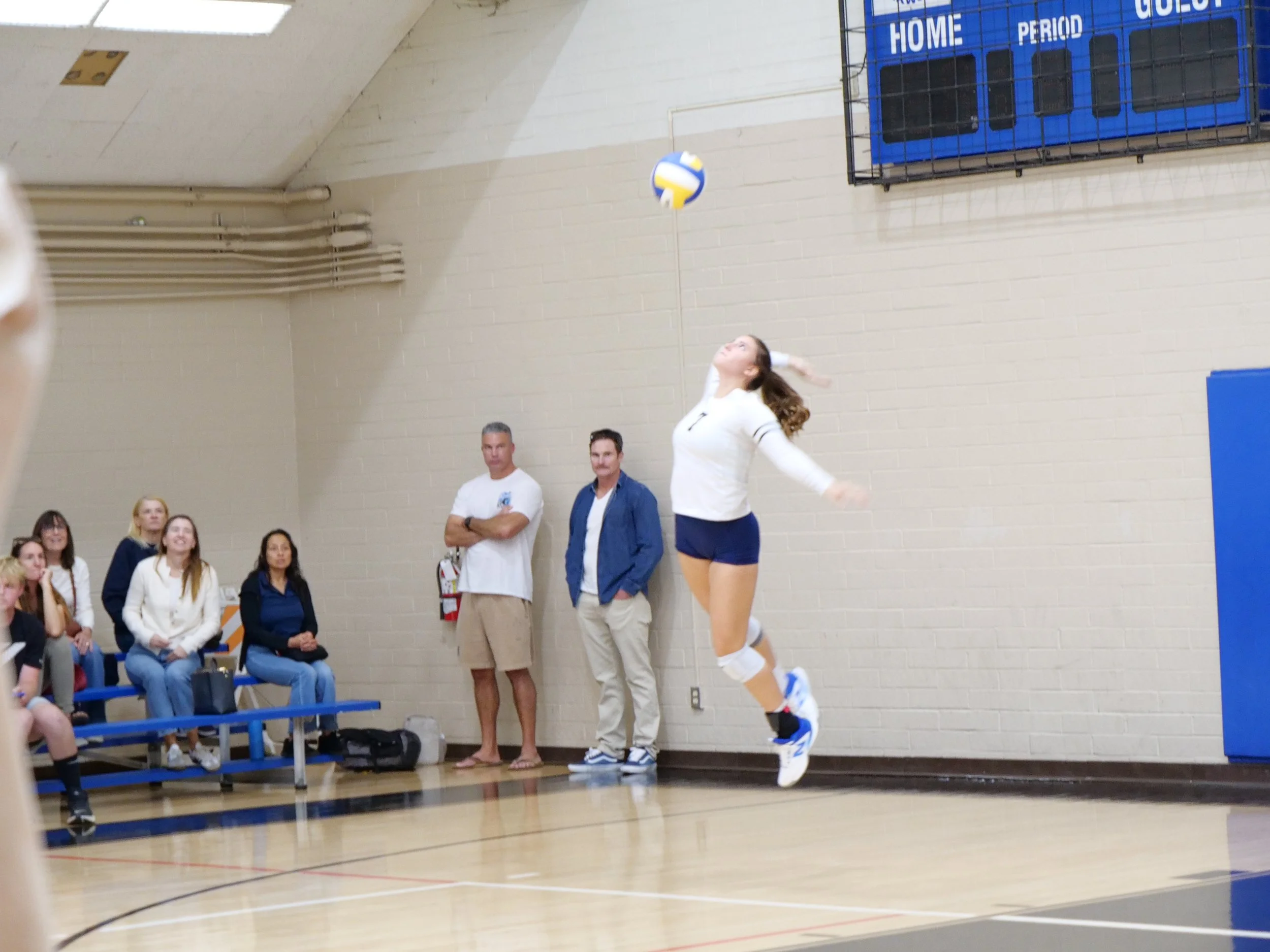 A volleyball player wearing a white jersey and shorts jumps to hit a volleyball in a gymnasium, with several spectators and officials watching.