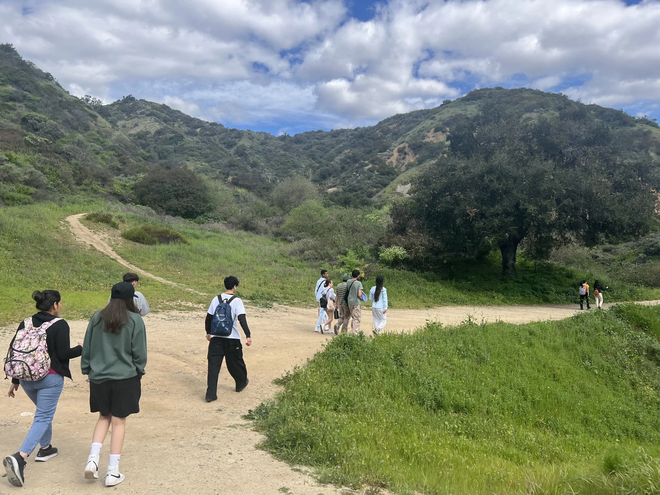 Group of people walking on a trail through a lush green mountainous area with trees and cloudy sky overhead.
