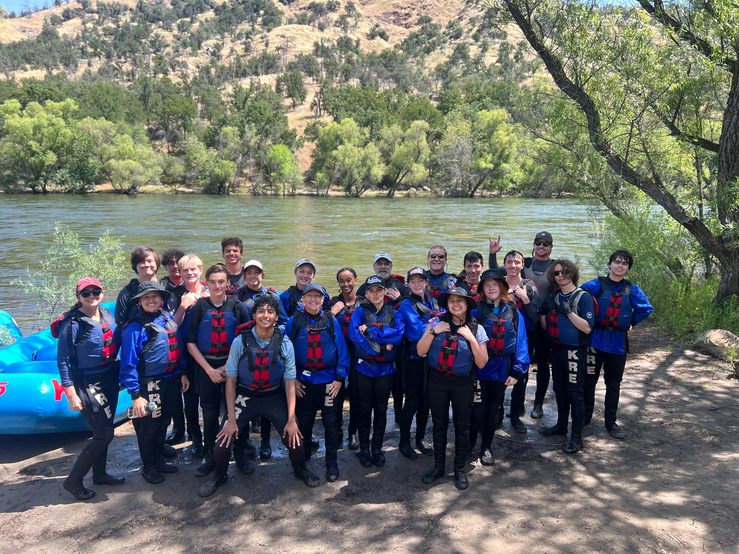 A group of about thirty people, including teenagers and adults, are standing outdoors near a river with hills and trees in the background. They are dressed in outdoor gear, some wearing life jackets, and appear to be on a rafting or kayaking trip. Th