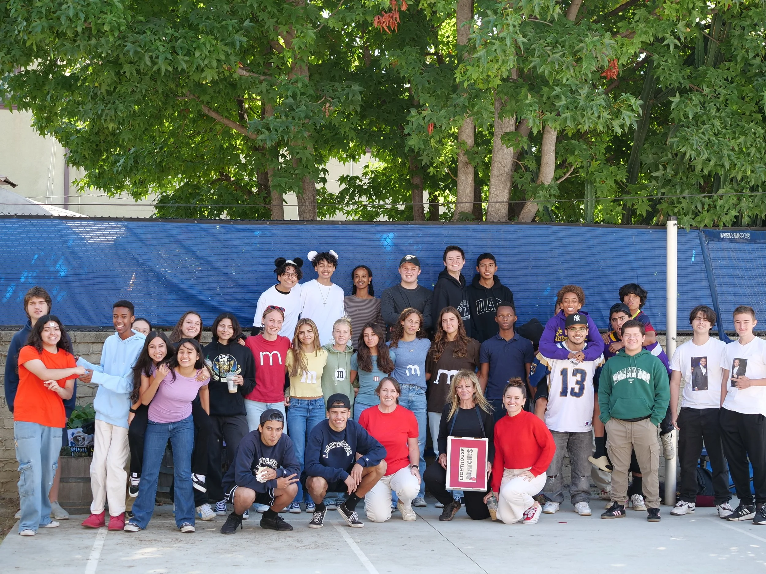A large group of diverse young people posing together outdoors under green trees, with a blue fence in the background.