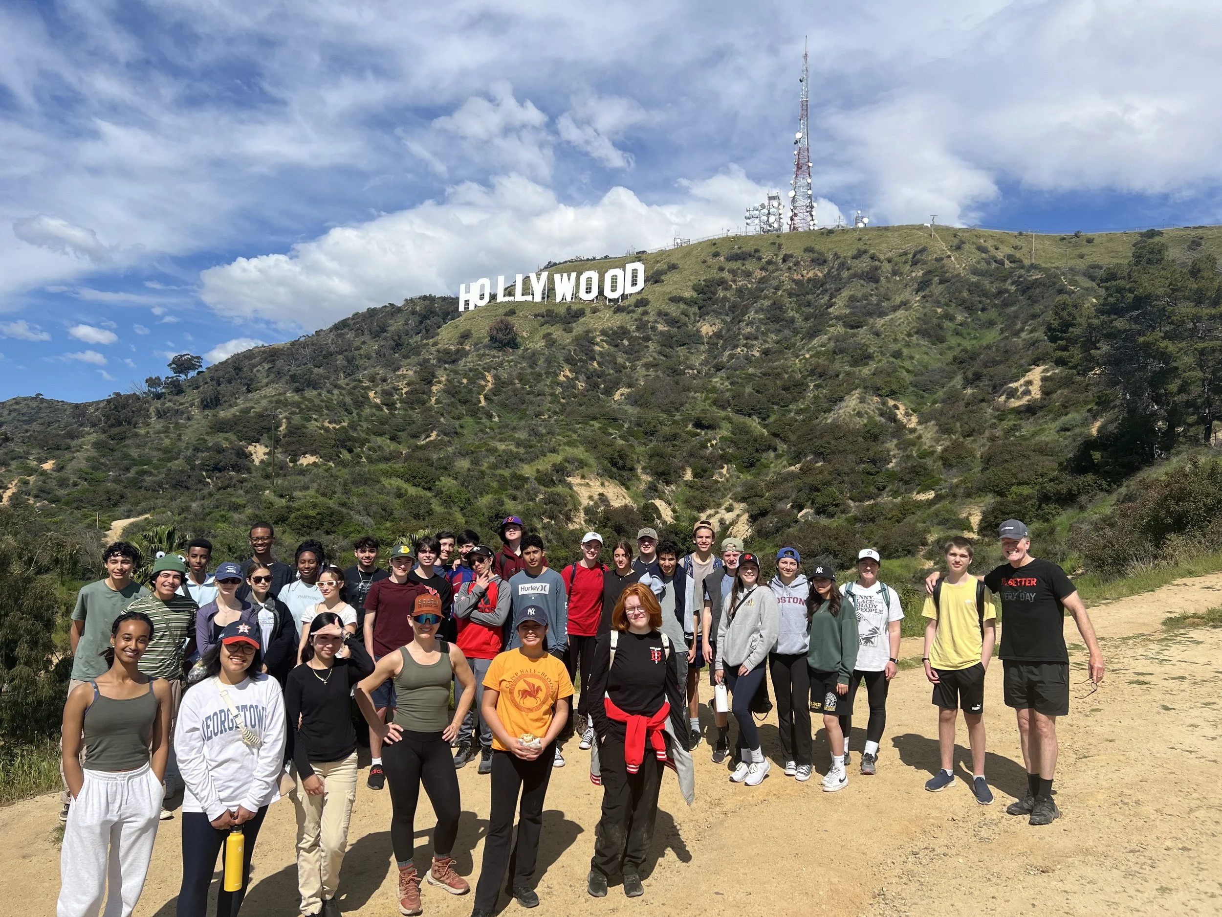 Group of young people posing for a photo on a dirt trail with the Hollywood sign visible on a hillside in the background under a partly cloudy sky.