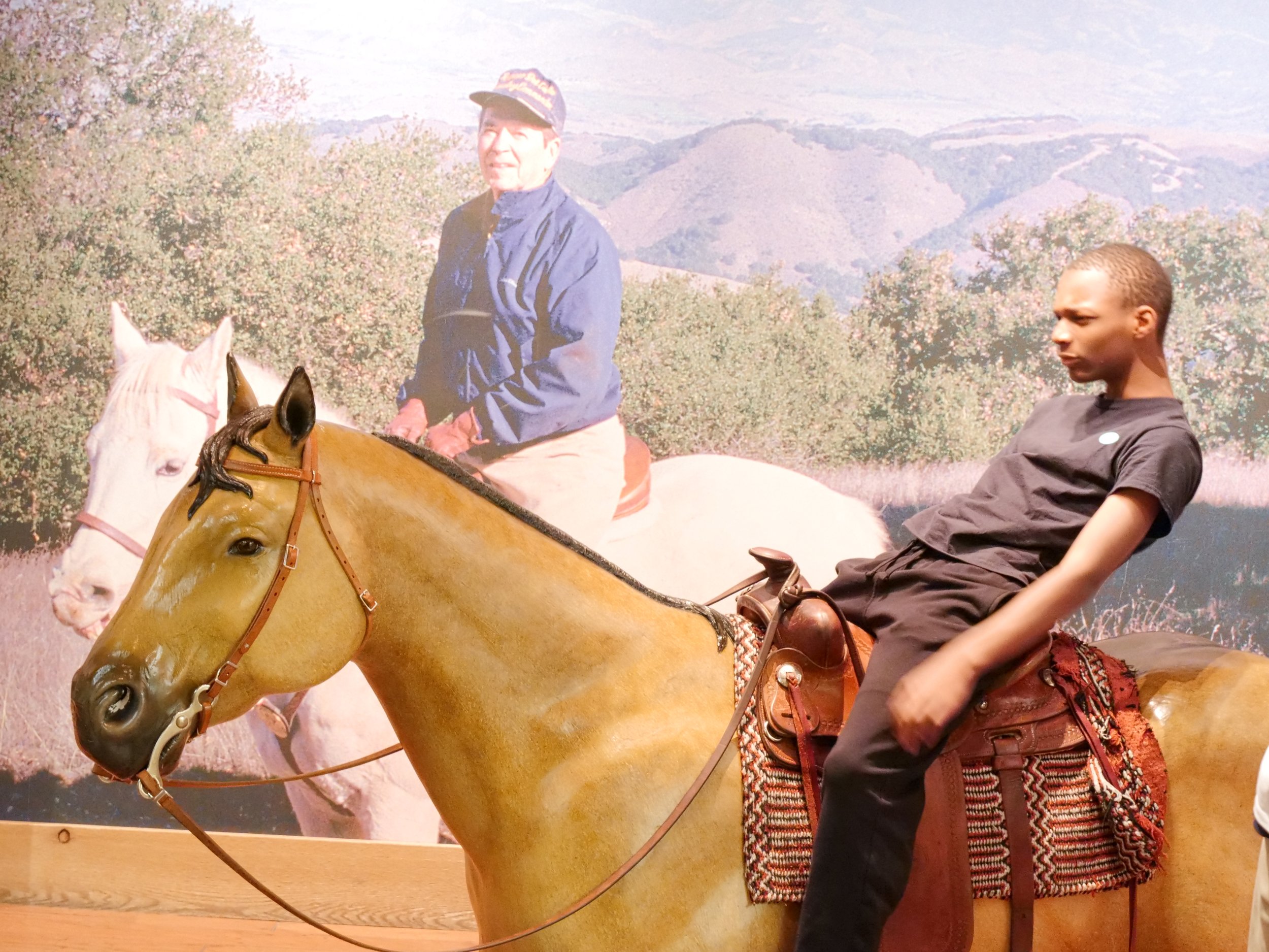 A person riding a white horse with a pink bridle in an outdoor setting with mountains and trees in the background. A young individual with short hair wearing a black shirt is sitting on the horse, and an older person wearing a blue jacket and a cap i