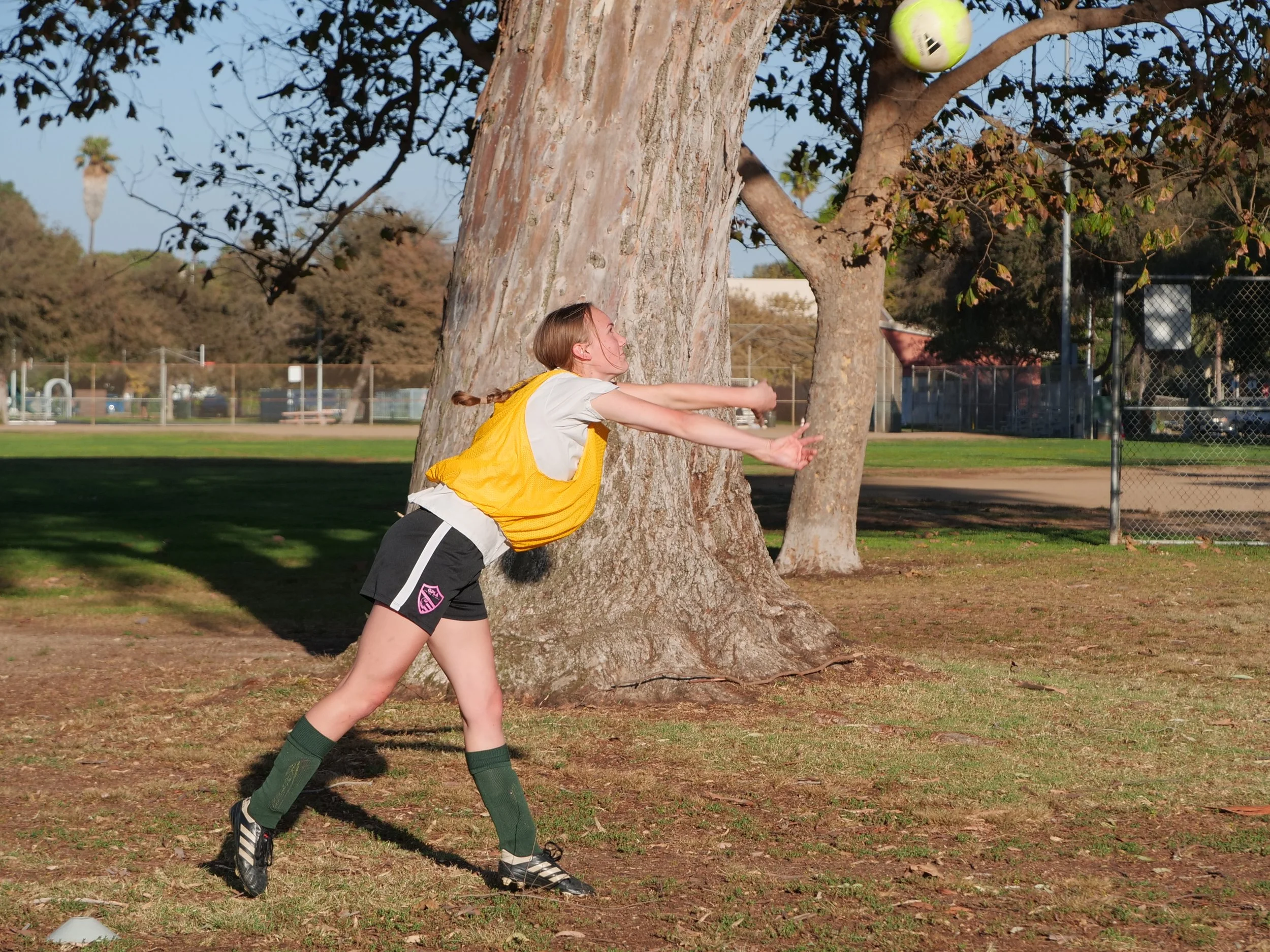 A young girl playing soccer outdoors, kicking a ball with her arms stretched forward and leaning against a tree, with a tennis court and a chain-link fence visible in the background.
