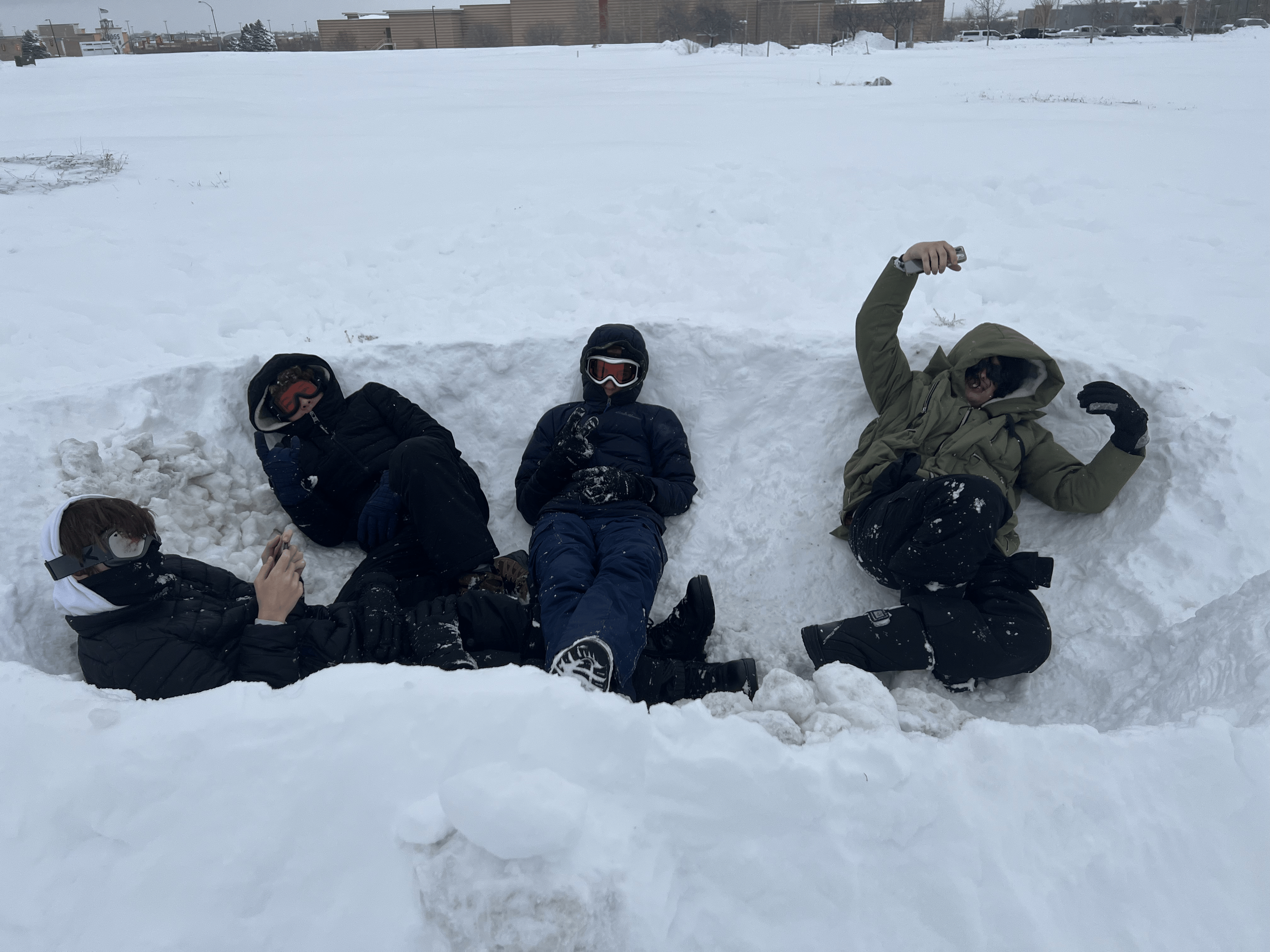 Four people sitting inside a snow fort in a snowy outdoor area during winter. One person is wearing goggles, a blue jacket, and gloves, while another is in a brown jacket and gloves. The other two are partially visible, with one using a phone and the