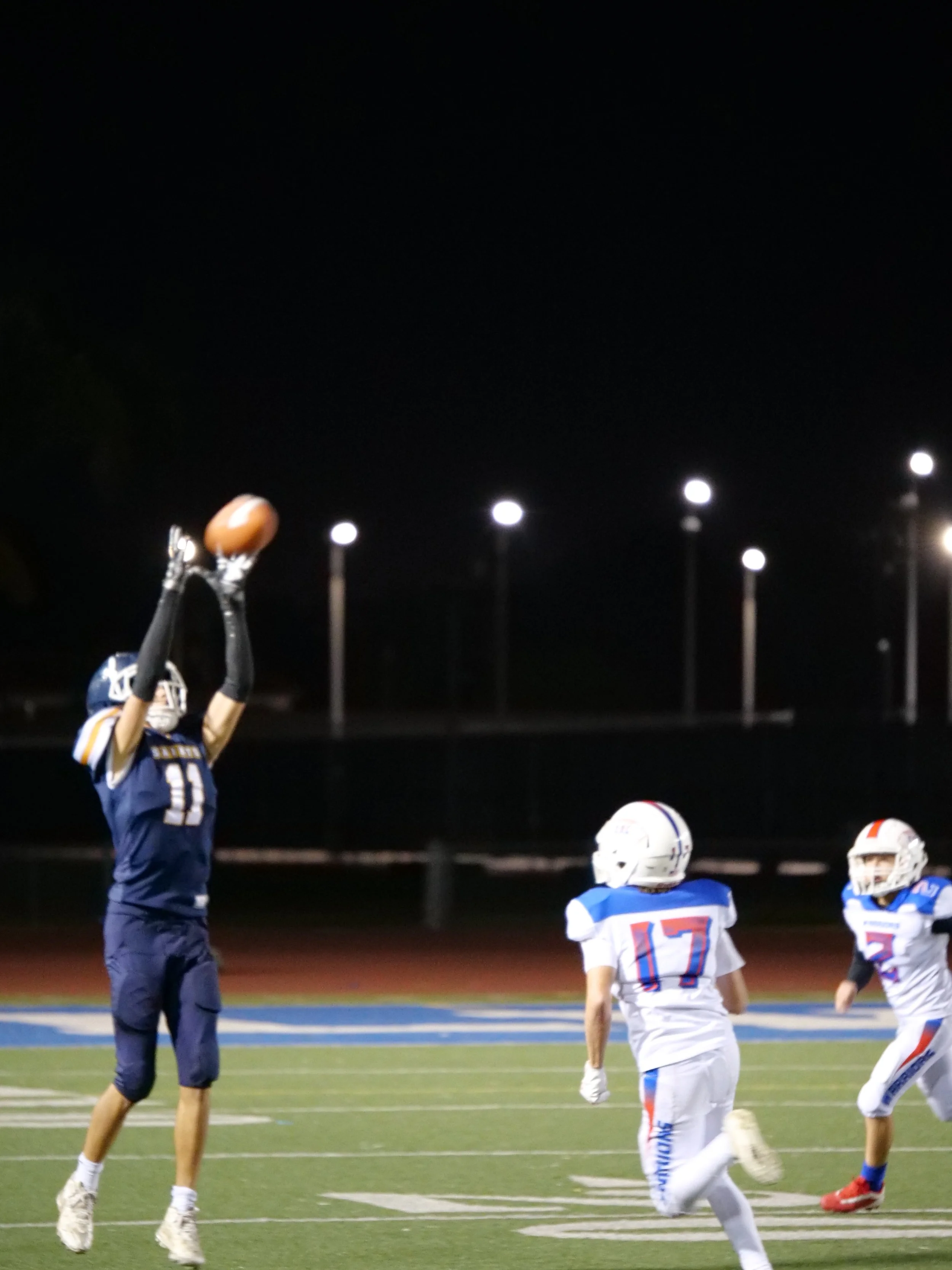 A football game at night showing a player in a dark uniform jumping to catch the football, with two players from the opposing team in white uniforms on the field.