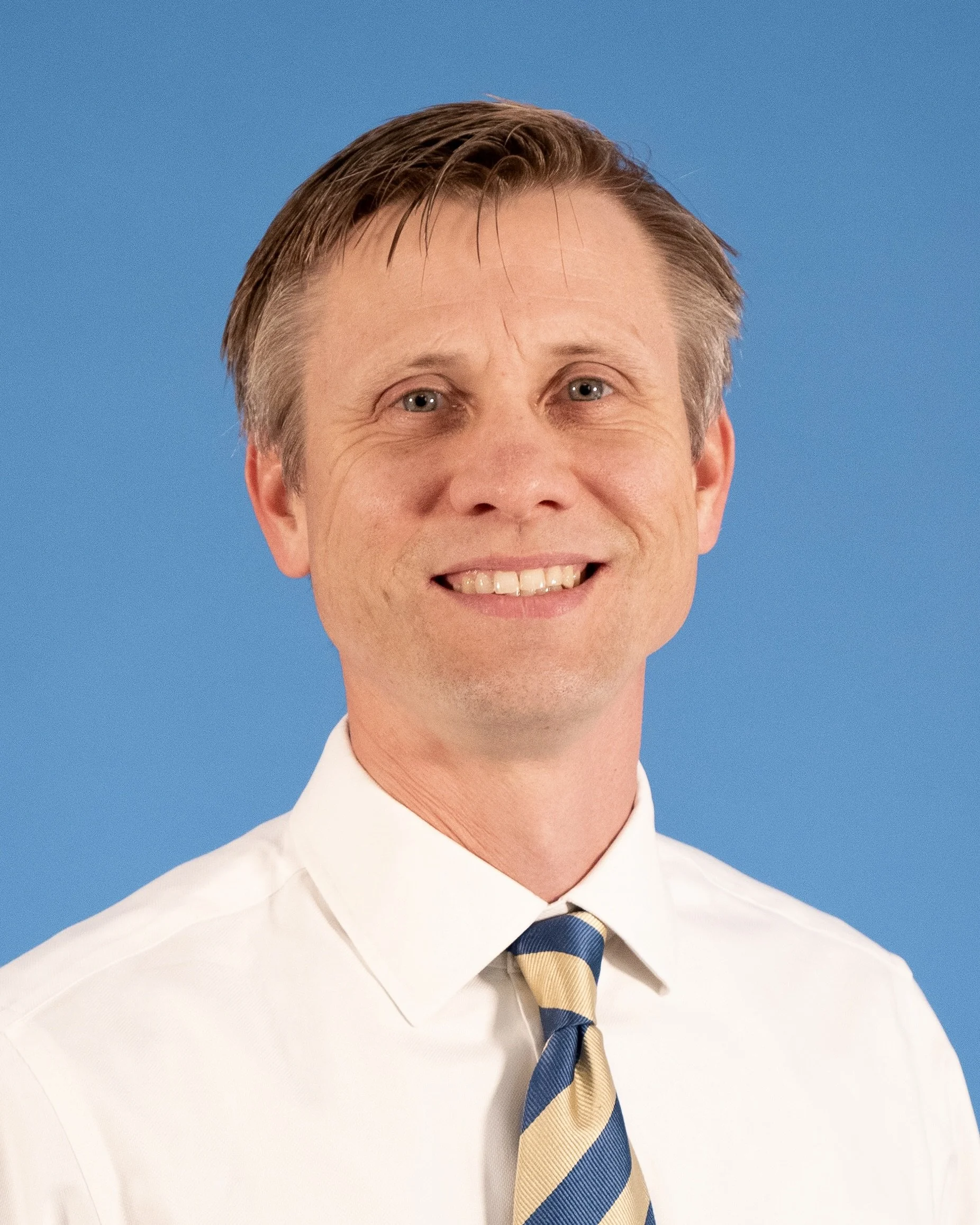 Smiling man in a white shirt and striped blue and white tie against blue background.