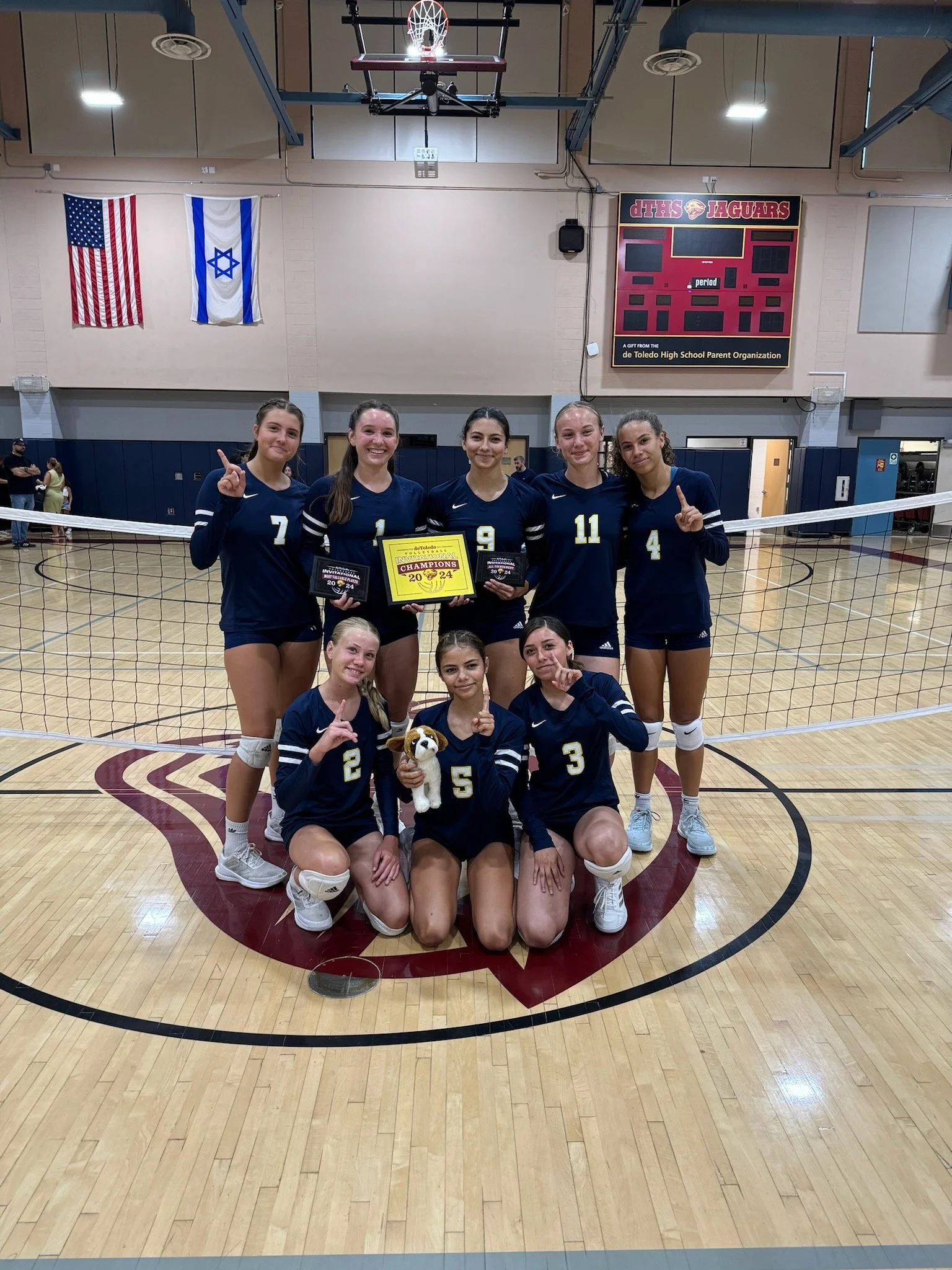 Girls volleyball team celebrating victory on an indoor court, holding awards and a trophy, smiling, with a volleyball net in the foreground.